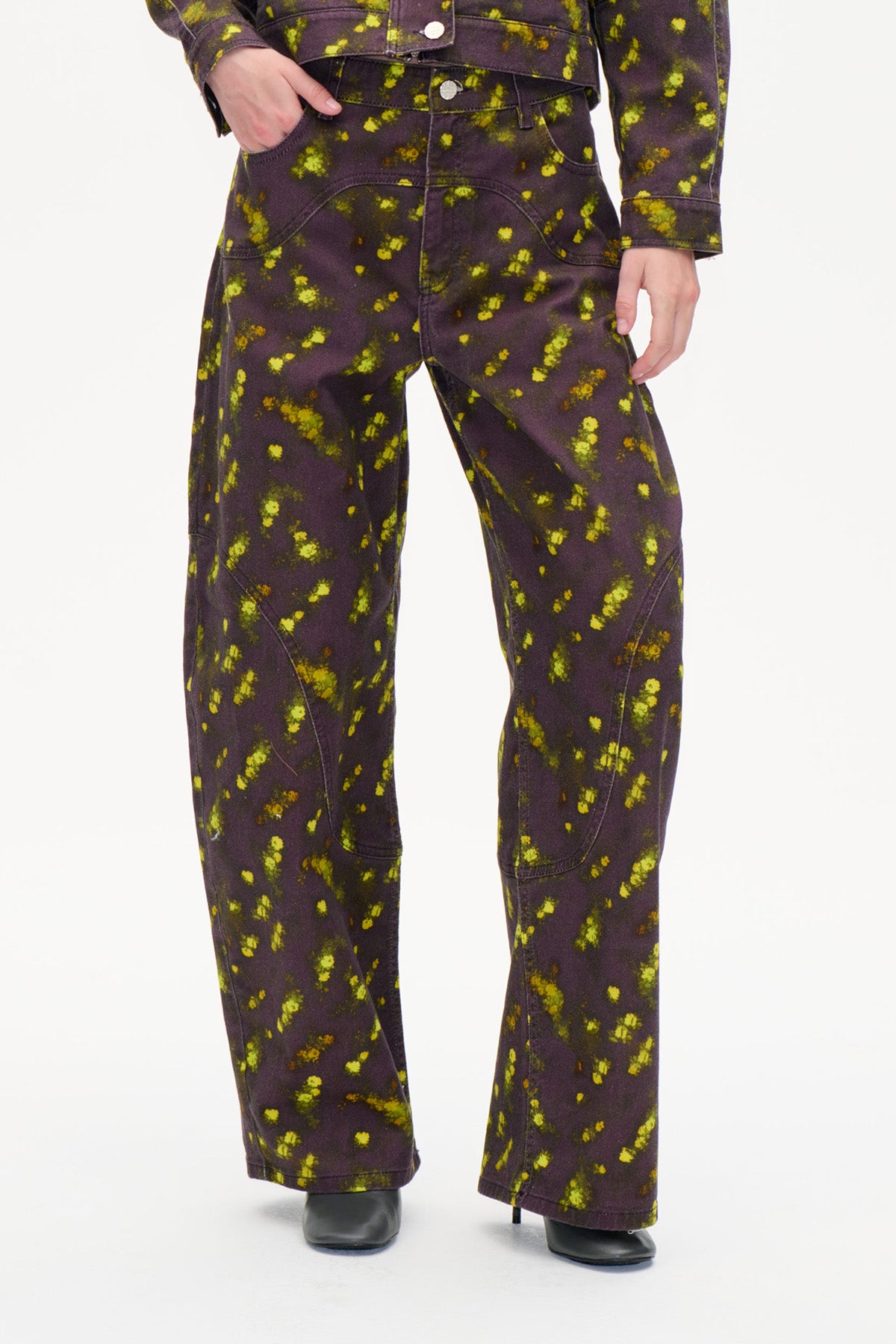 Detailaufnahme der Vorderseite einer weiten, dunkelpflaumenfarbenen Hose mit leuchtend gelb‑grünem Sprenkel-Print und geschwungenen Teilungsnähten.