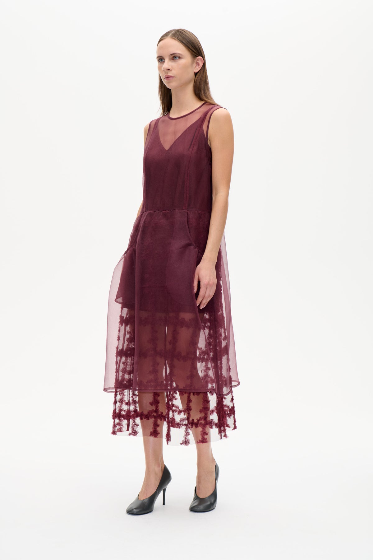 Model wearing a plum sleeveless dress with a sheer overlay and dark floral embroidery, shown from the front against a white background.