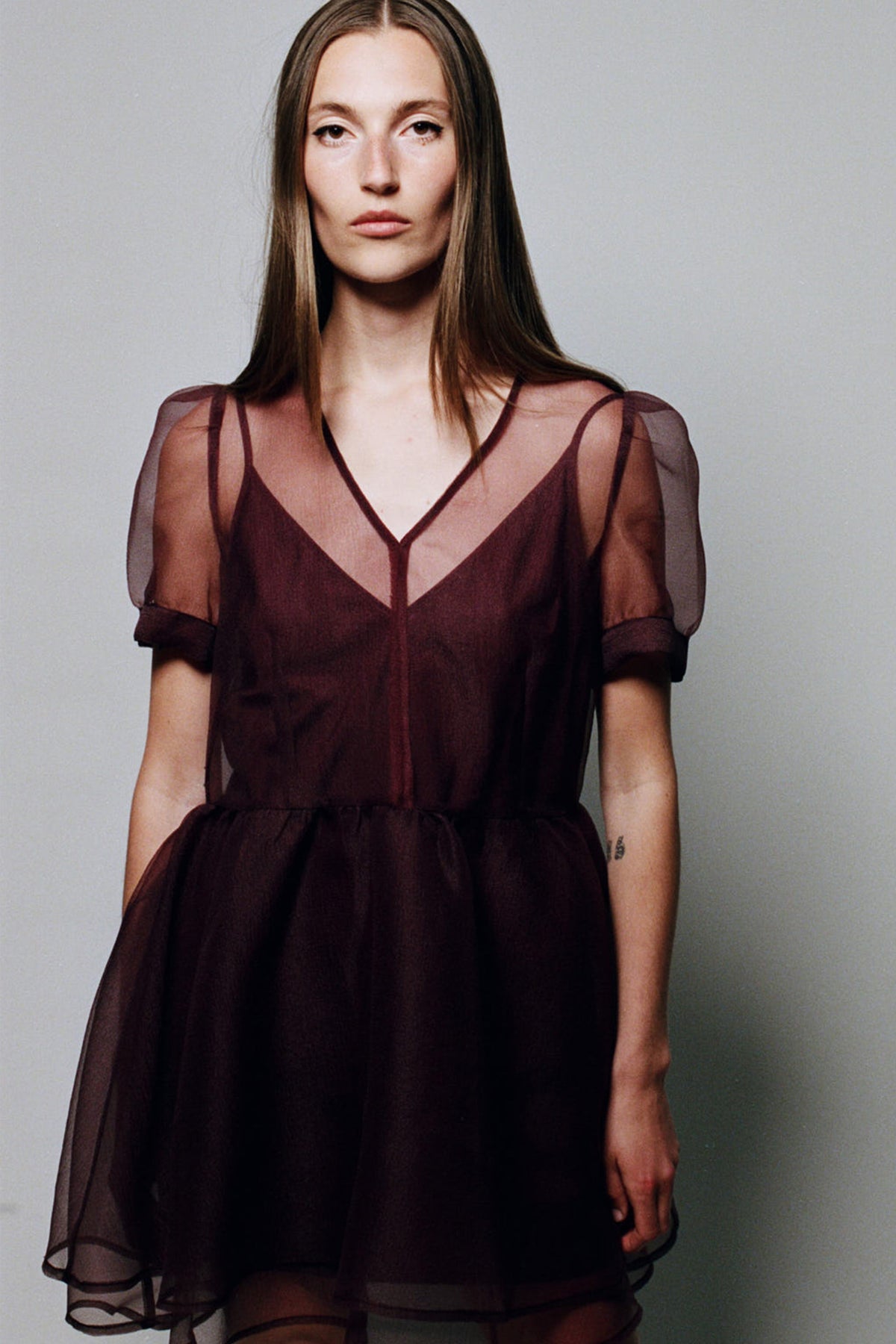 Frontal campaign portrait of a model wearing a dark burgundy dress with a V-neck, sheer short sleeves and a flowing sheer overlay.