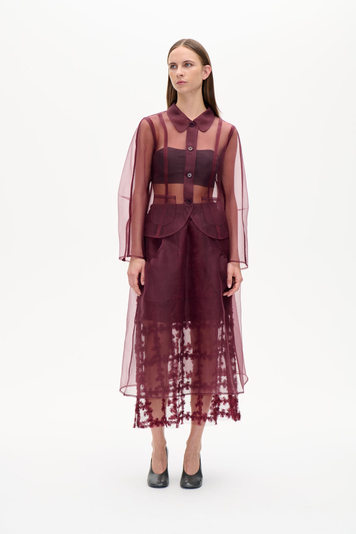 Model wearing a sheer plum button-front tunic with a rounded collar and vertical panels over a dark top and floral skirt.