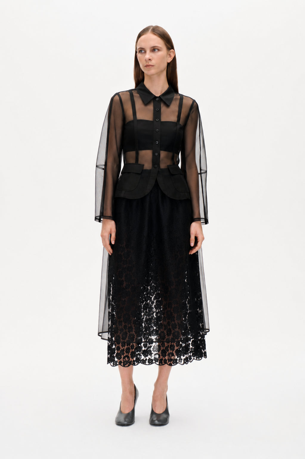 Front view of a sheer black button-front blouse with a pointed collar and flap pockets, worn over a black top with a long scalloped lace skirt.