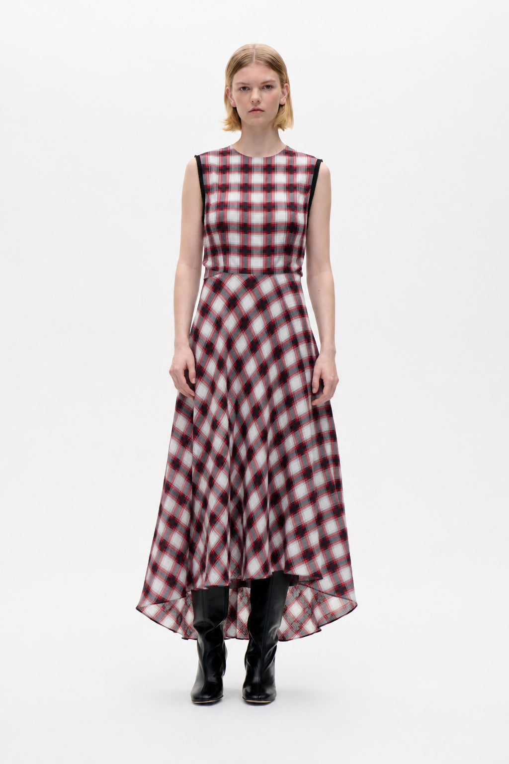 Model draagt een mouwloze geruite top in rood, zwart en grijs, een bijpassende maxi-rok met hogere voorkant en lagere achterkant en zwarte kniehoge laarzen.