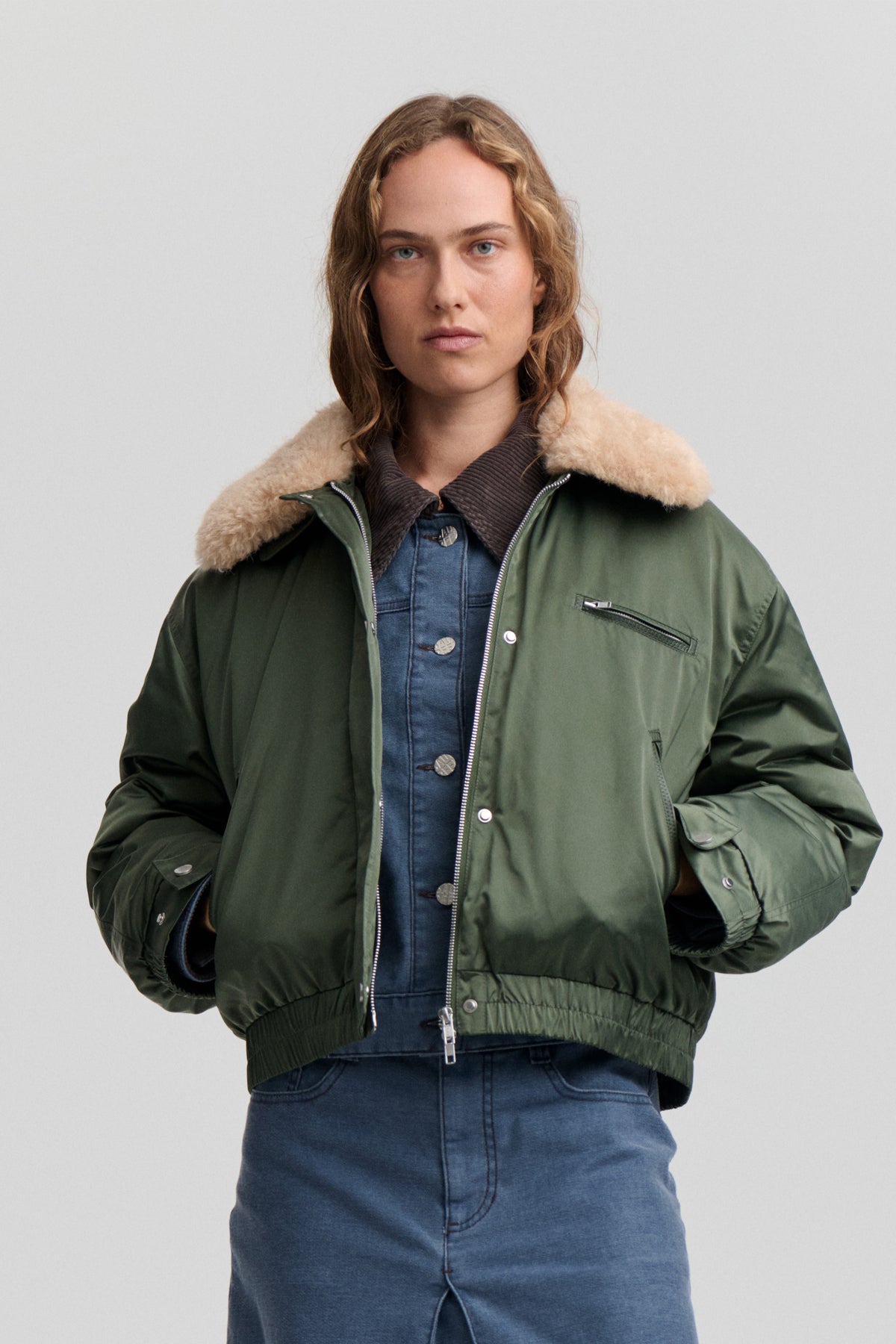 Front view of a model wearing a dark green zip-front jacket with a fluffy beige collar and chest pocket over blue denim.