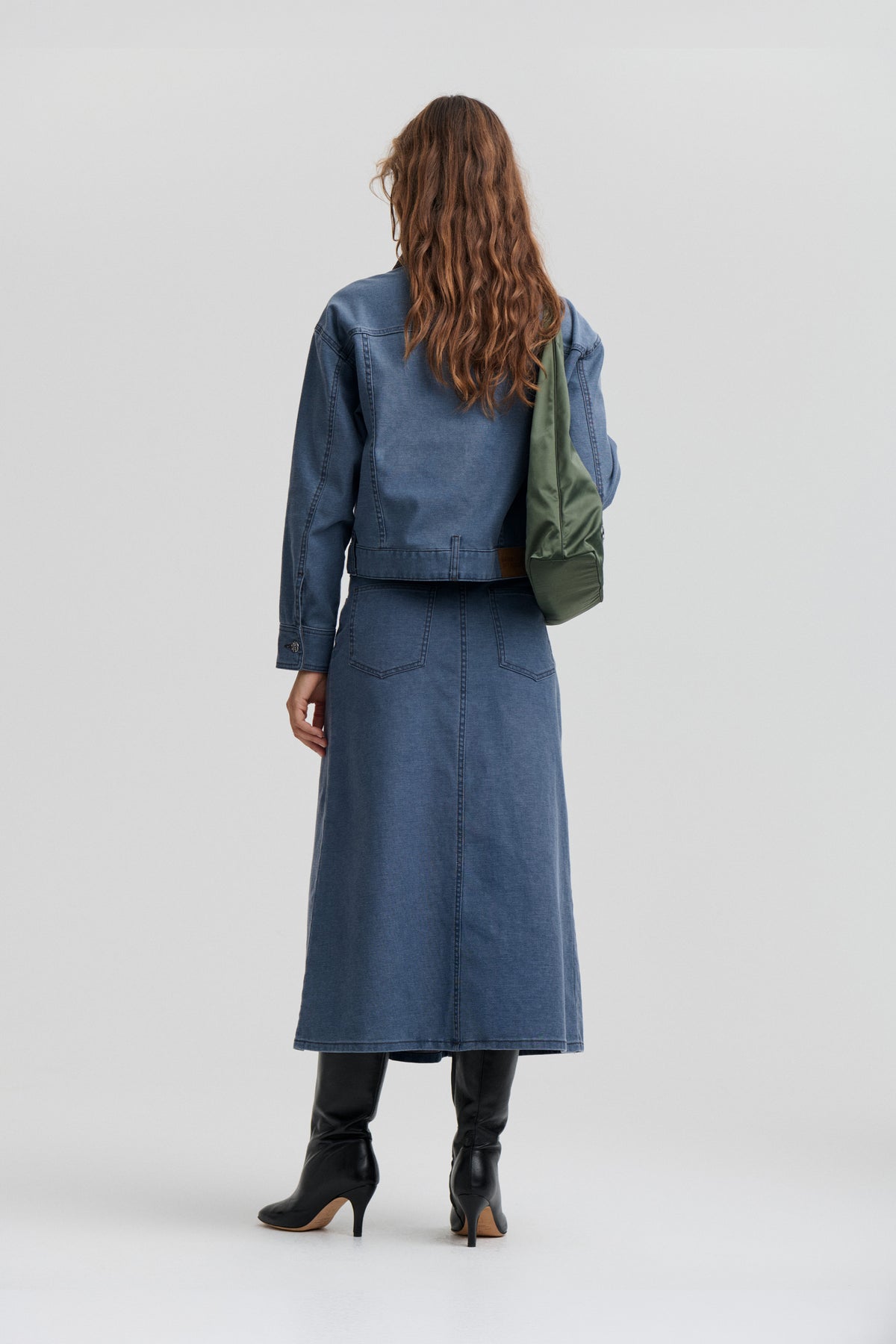 Back view of a model in a blue denim jacket and matching long skirt, carrying an olive-green shoulder bag and wearing black heeled boots.