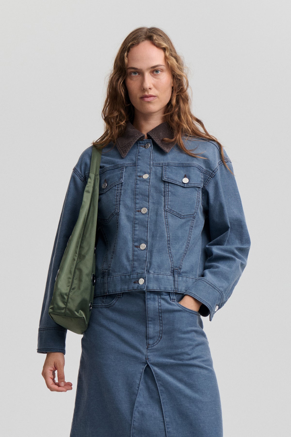 Model wearing a blue denim jacket with a dark corduroy collar and buttoned chest pockets, paired with a matching denim skirt and olive shoulder bag