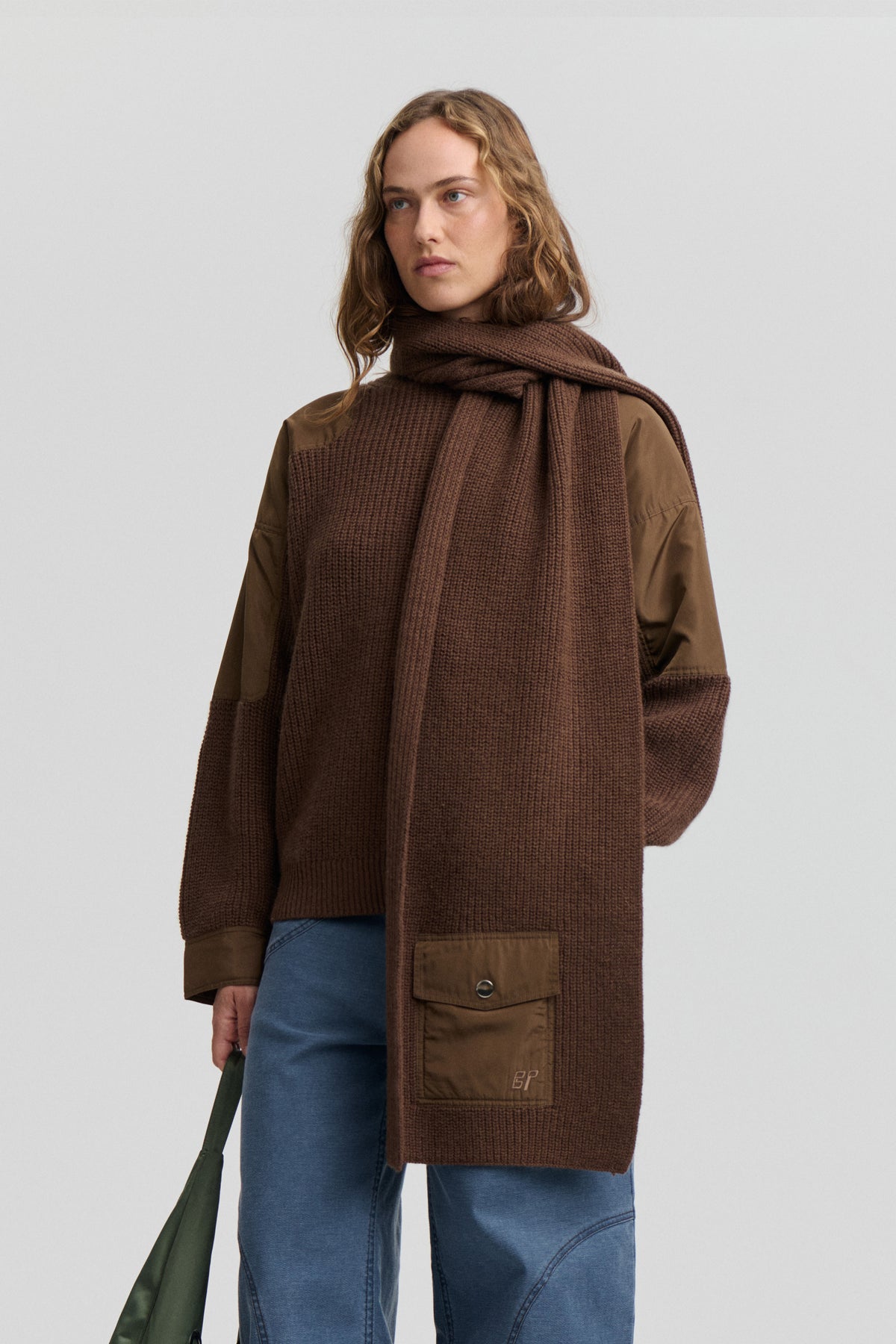 Model wearing a long, textured dark brown knit scarf wrapped around the neck over an olive jacket and blue jeans.