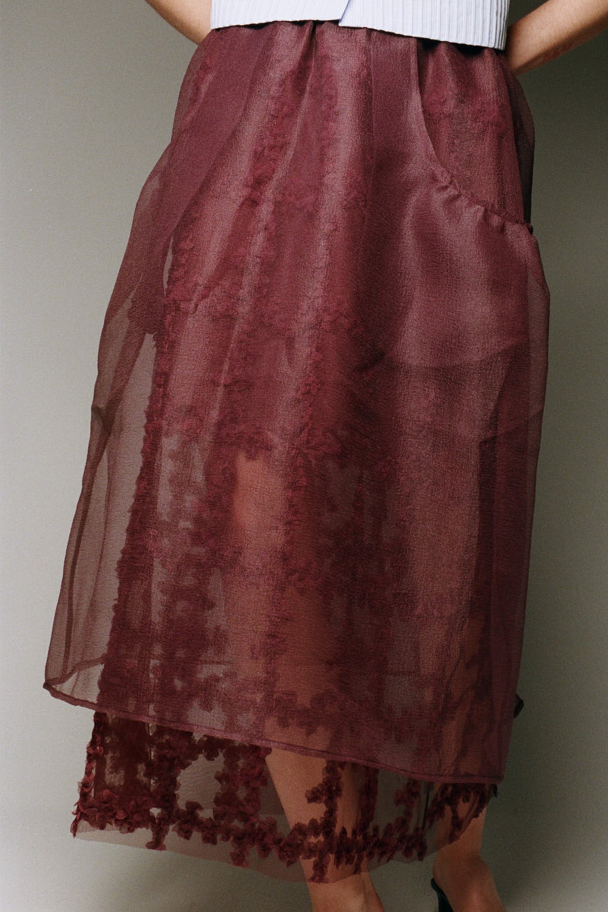 Close-up campaign view of a model wearing a sheer burgundy gathered skirt with a floral lace-trimmed hem and a white ribbed top.