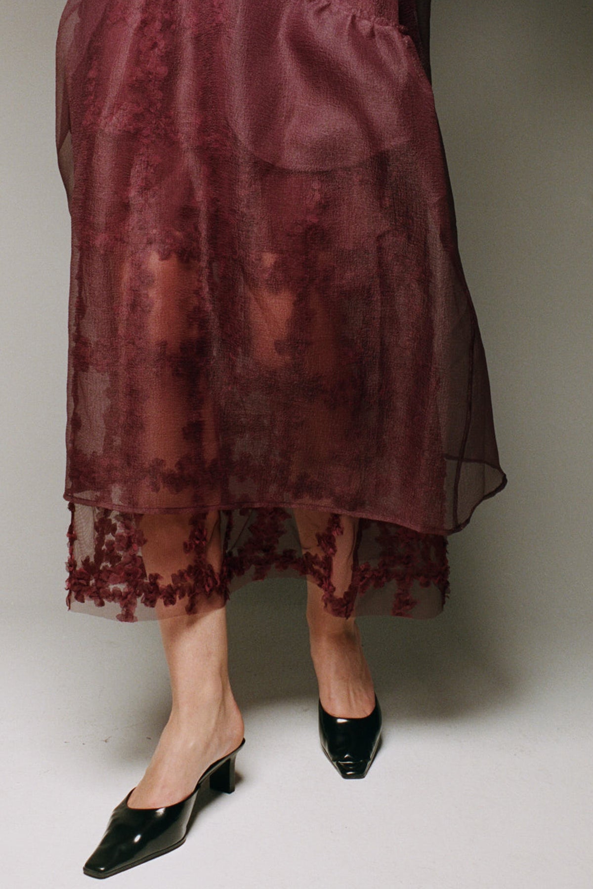 Close-cropped campaign view of a model wearing a sheer burgundy skirt layered over a floral lace hem, with black pointed shoes.
