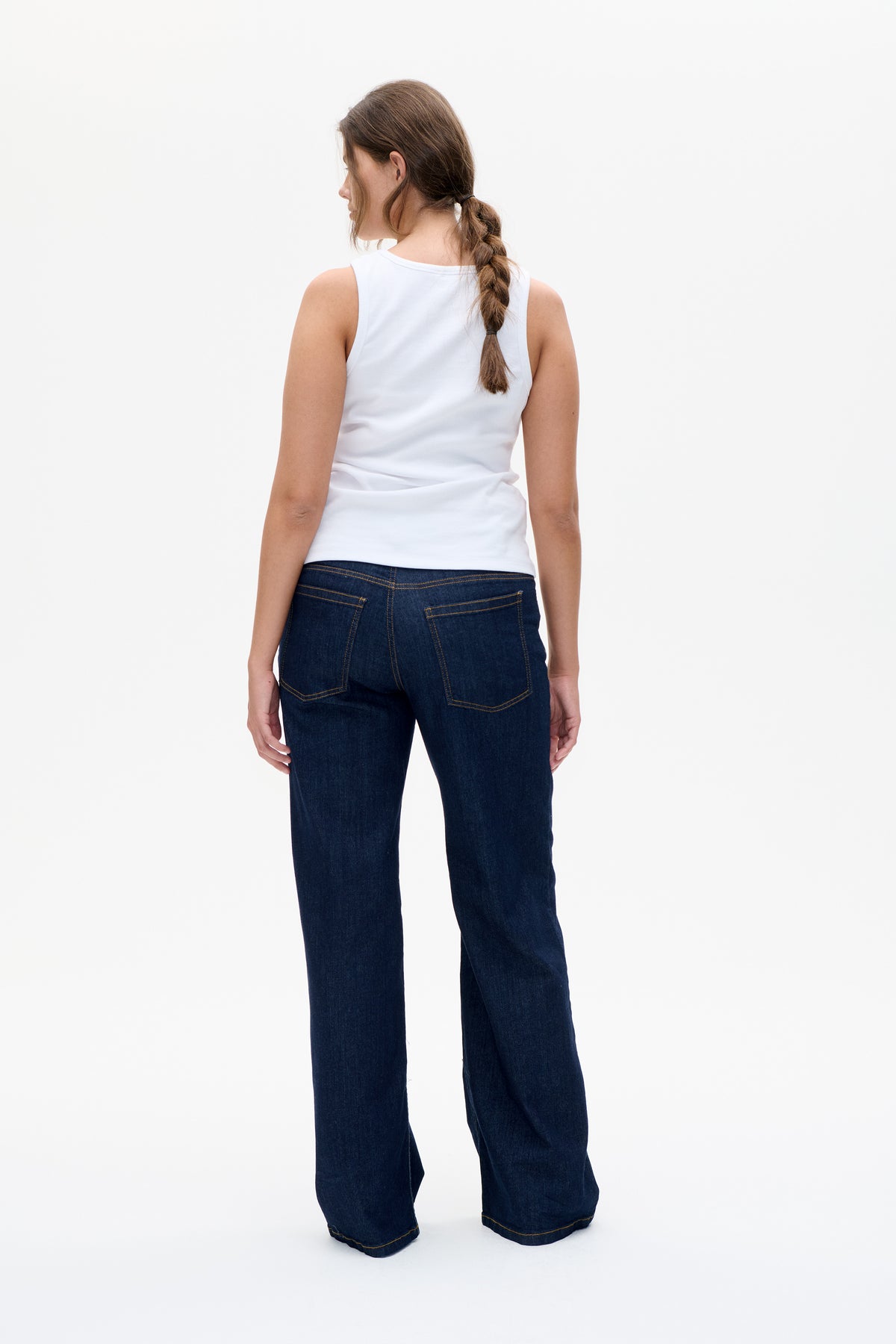 Back view of a model wearing a white sleeveless top and dark indigo wide-leg jeans with contrast stitching and patch pockets.