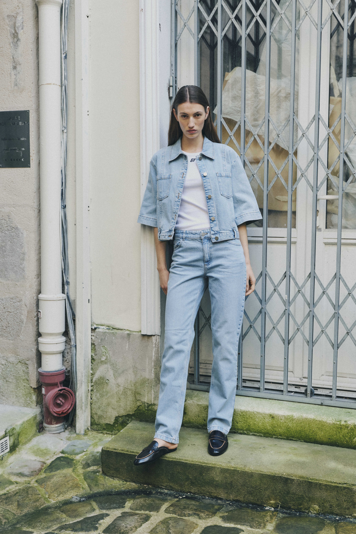 A model wears a light-wash denim jacket and matching jeans over a white graphic T-shirt, standing by a metal storefront gate.