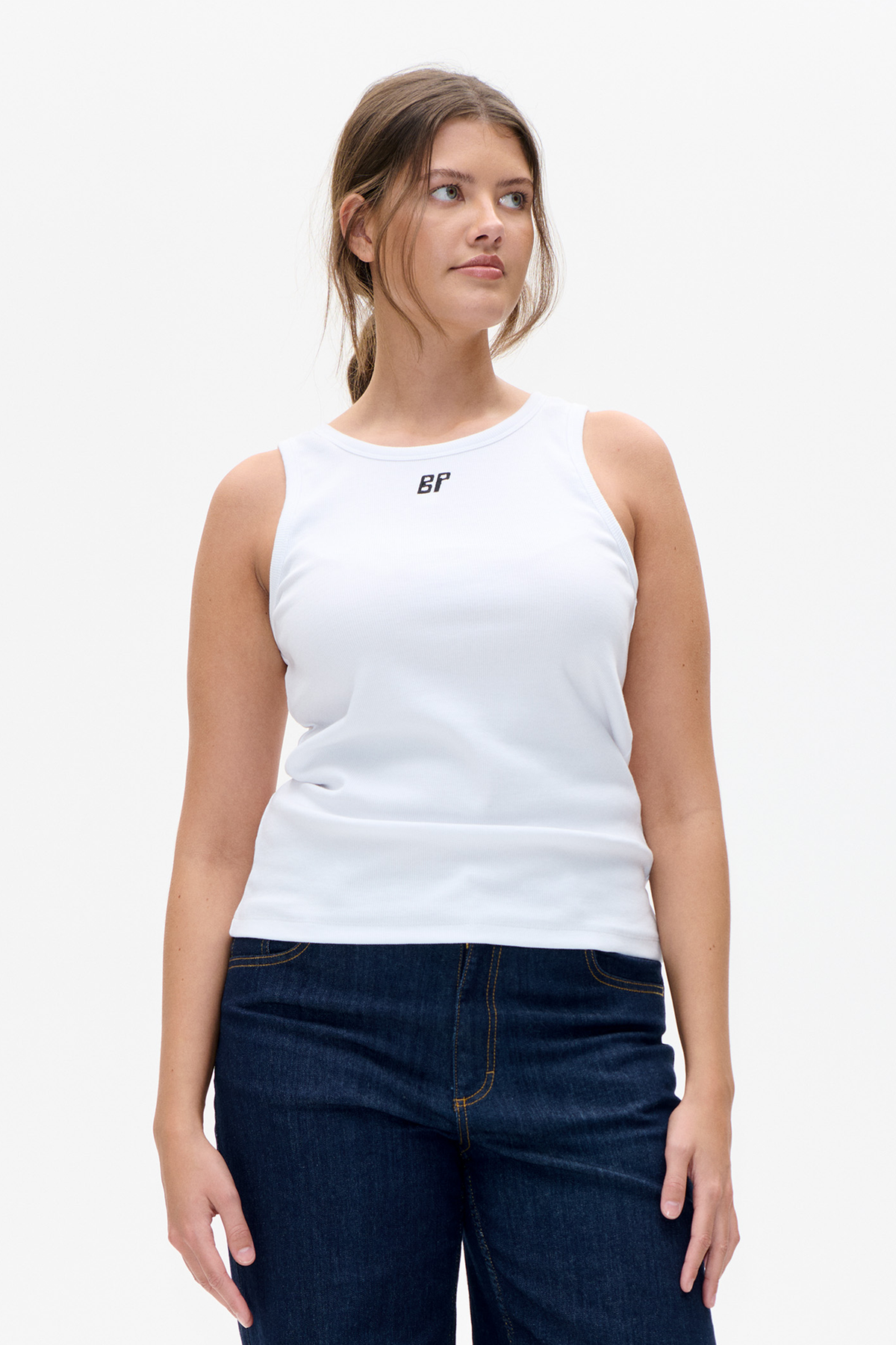 Model wearing a white sleeveless ribbed top with a small black chest logo and dark indigo jeans, shown from the front.