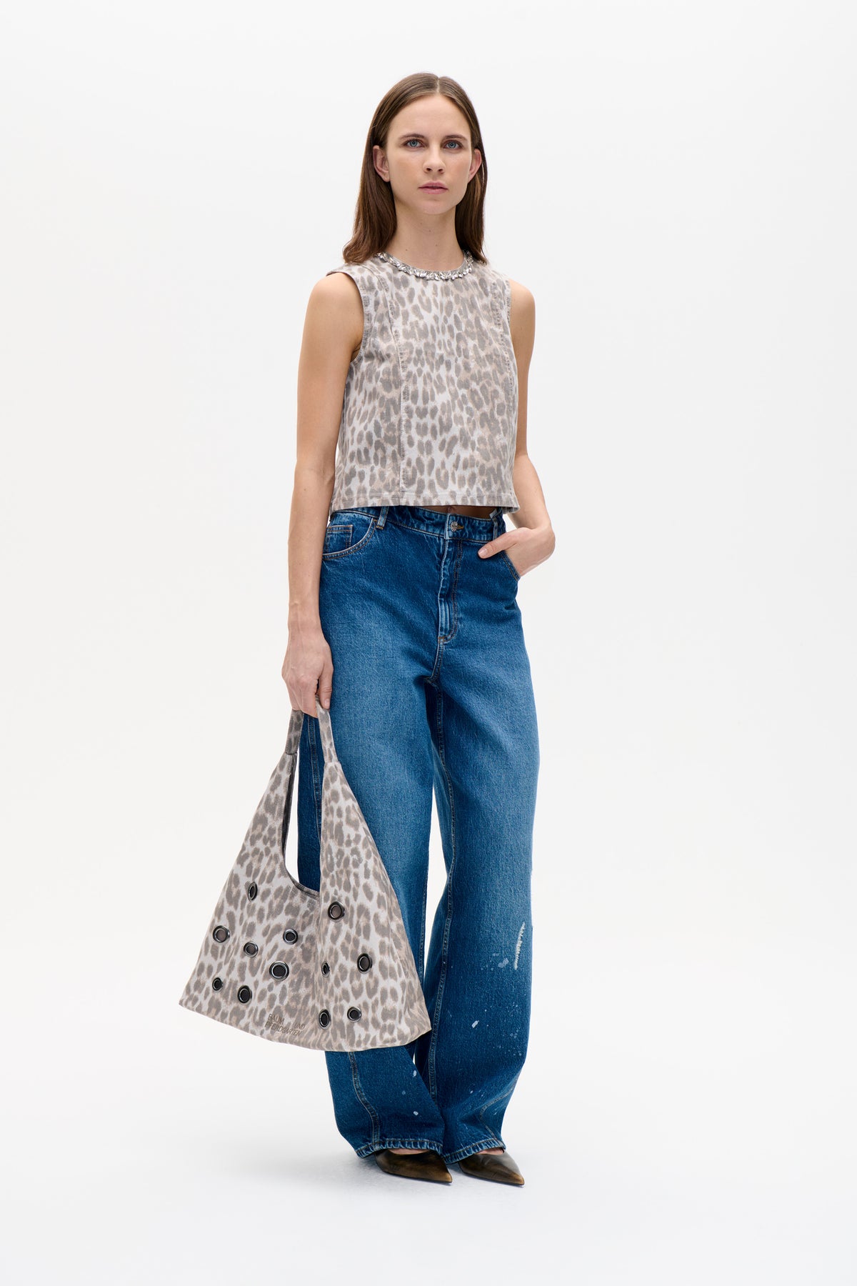Model wearing a sleeveless beige and grey animal-print top with a jeweled neckline, blue jeans, and a matching grommet-detail tote.