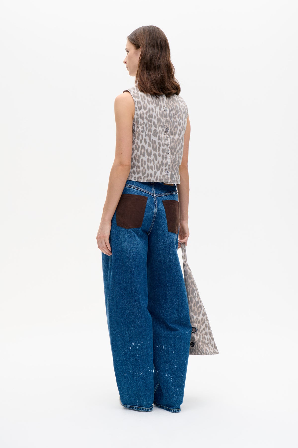 Model viewed from behind in a sleeveless beige and gray animal-print vest with silver back buttons and wide-leg blue jeans.