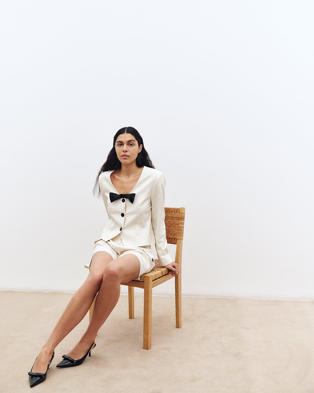 A seated model wears an ivory button-front jacket with a black bow, matching shorts and black pointed heels.
