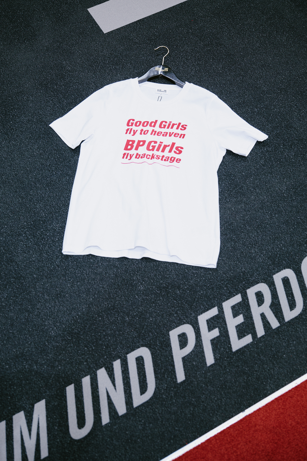 White short-sleeve T-shirt with pink text reading “Good Girls fly to heaven, BP Girls fly backstage,” hanging against a dark graphic backdrop.