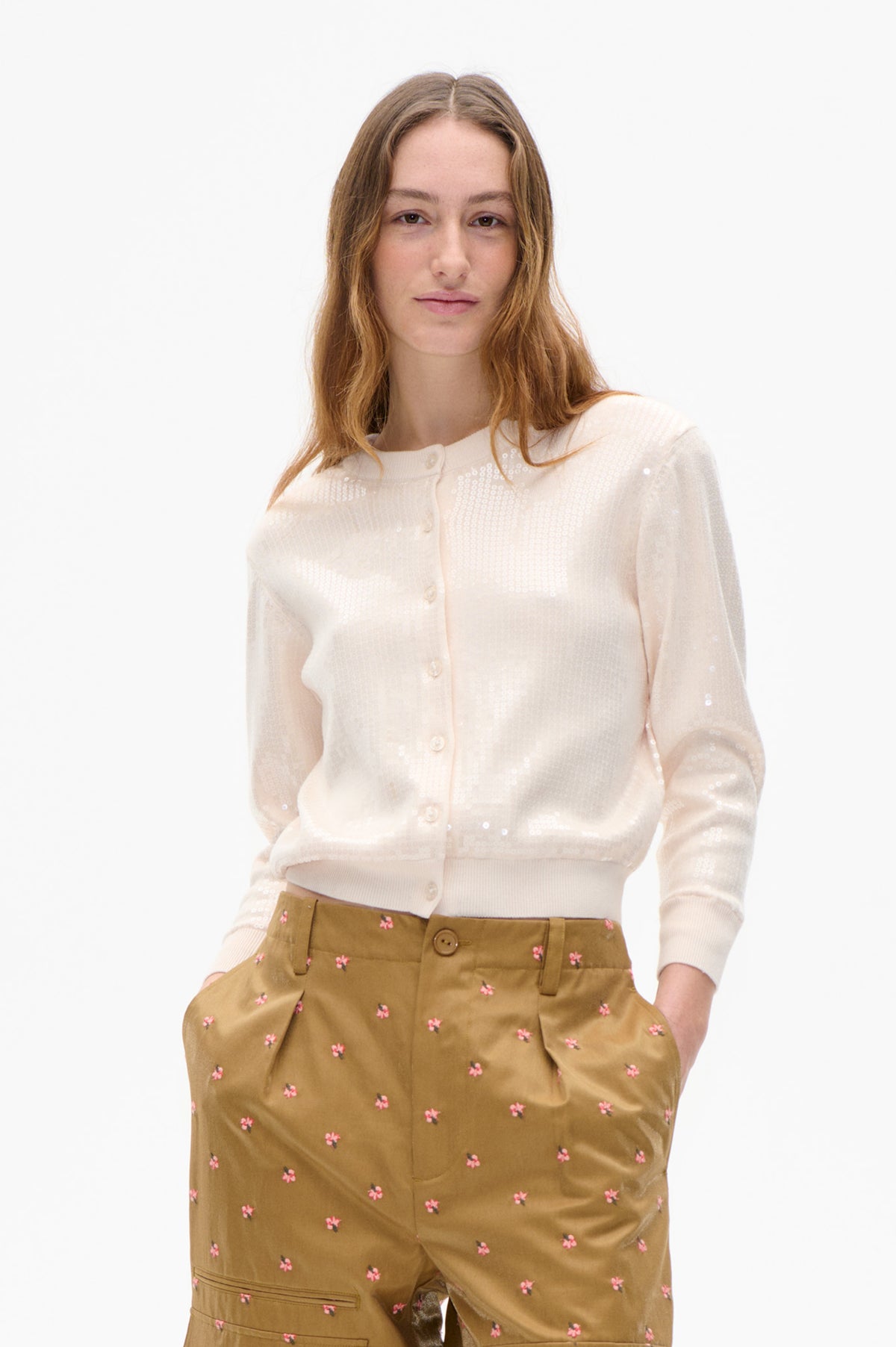 Model trägt eine cremefarbene Strickjacke mit Knopfleiste und dezenten Pailletten sowie eine braune Hose mit verstreuten kleinen rosa Blumen