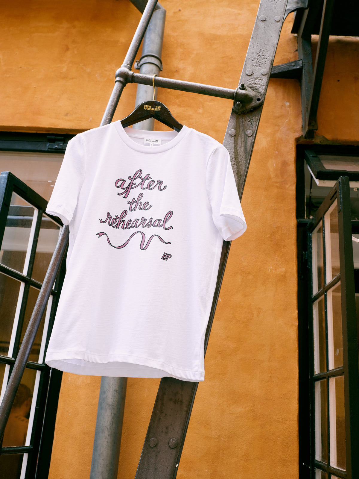 White short-sleeved T-shirt hanging on a black hanger, with pink and dark outlined script reading “After the rehearsal” on the front.