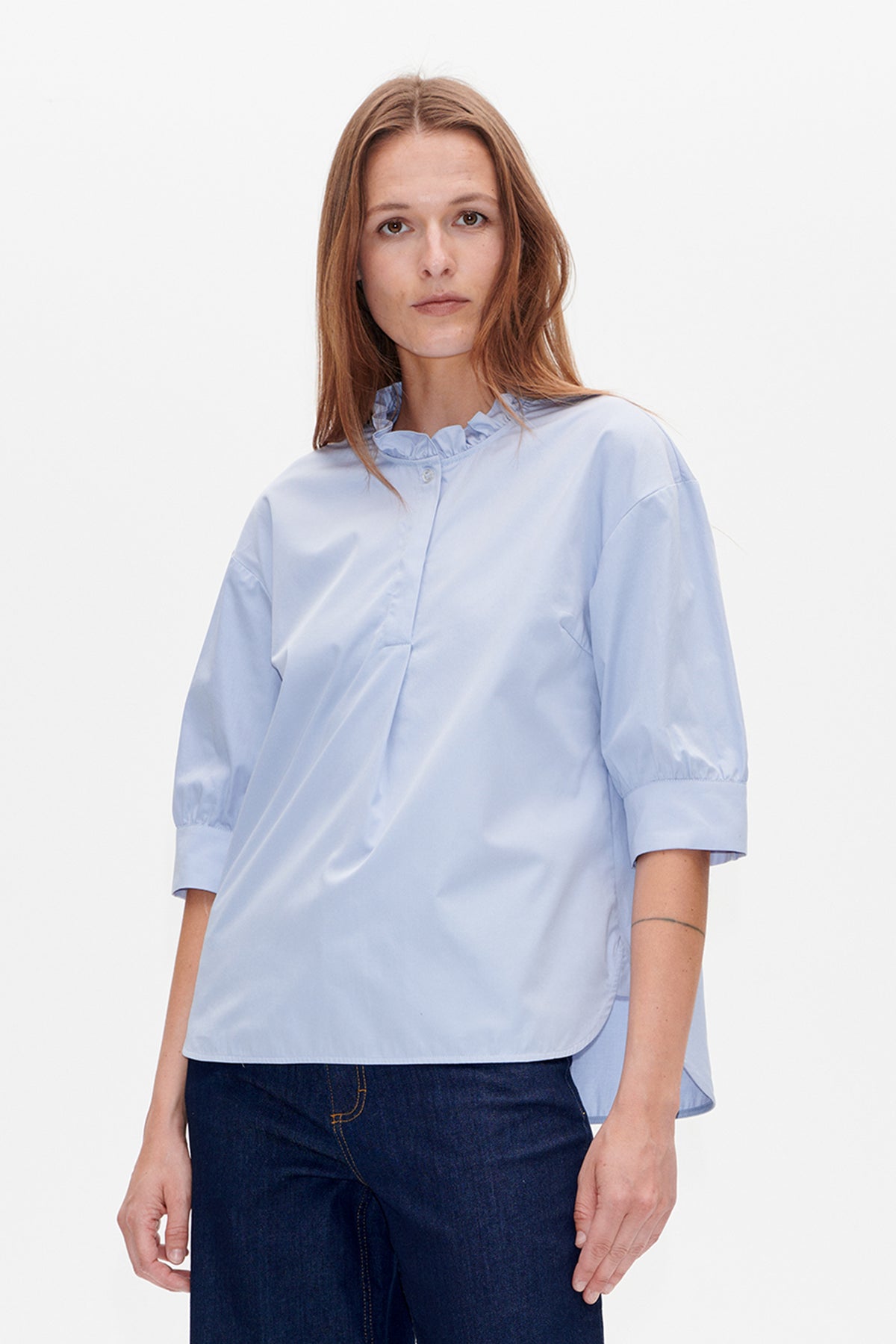 Front view of a pale blue blouse with a ruffled collar, button placket and elbow-length sleeves, worn with dark jeans.