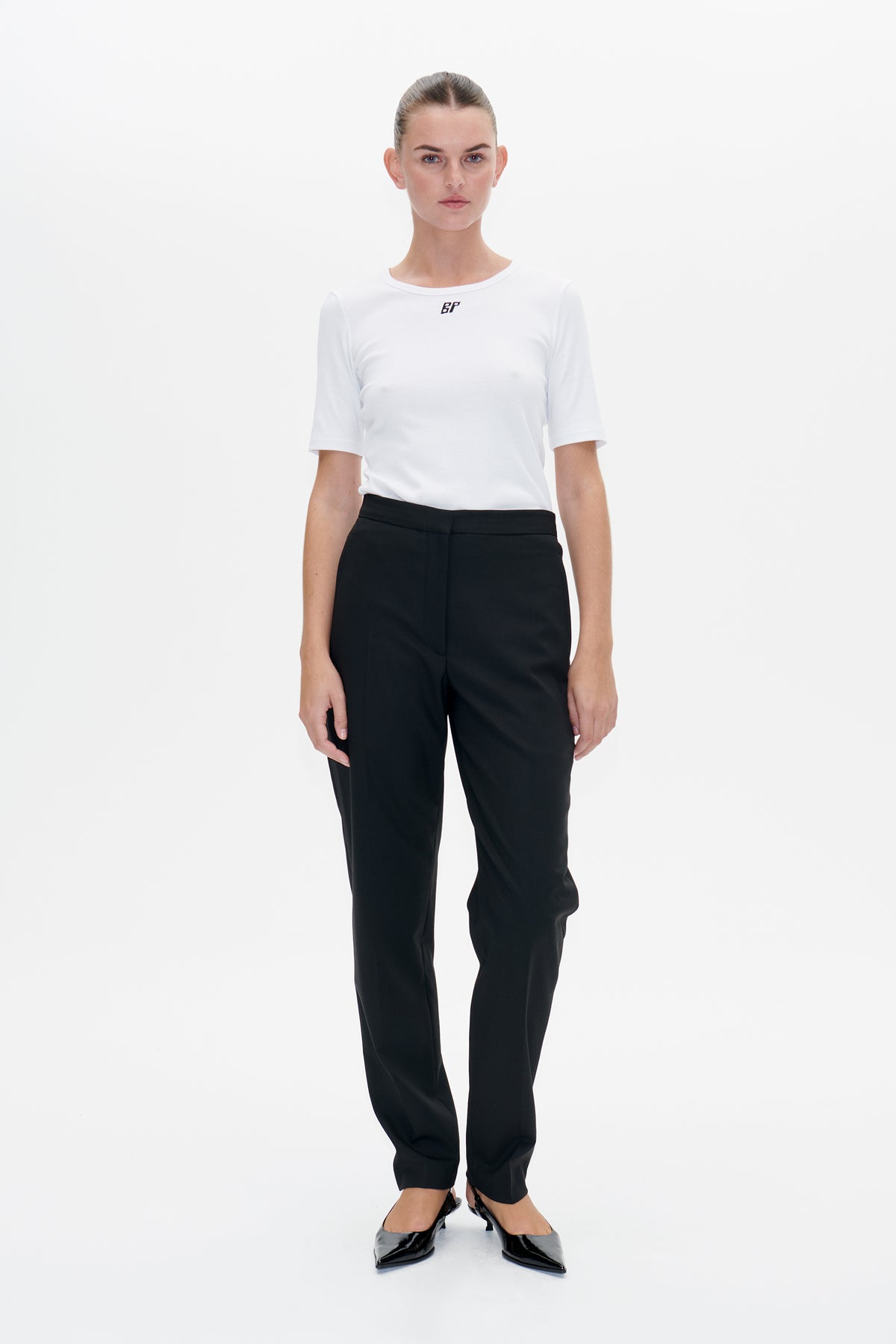 Vooraanzicht van een model met een wit T-shirt met korte mouwen en klein zwart borstlogo, een zwarte broek en puntige schoenen.