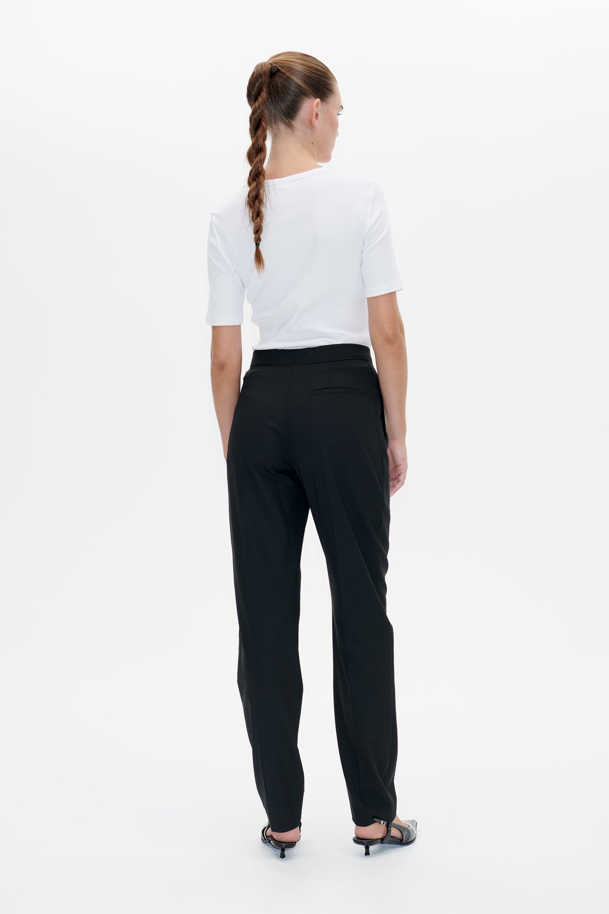 Achteraanzicht van een model dat een effen witte top met korte mouwen en een zwarte, getailleerde broek met een paspelzak aan de achterkant draagt.