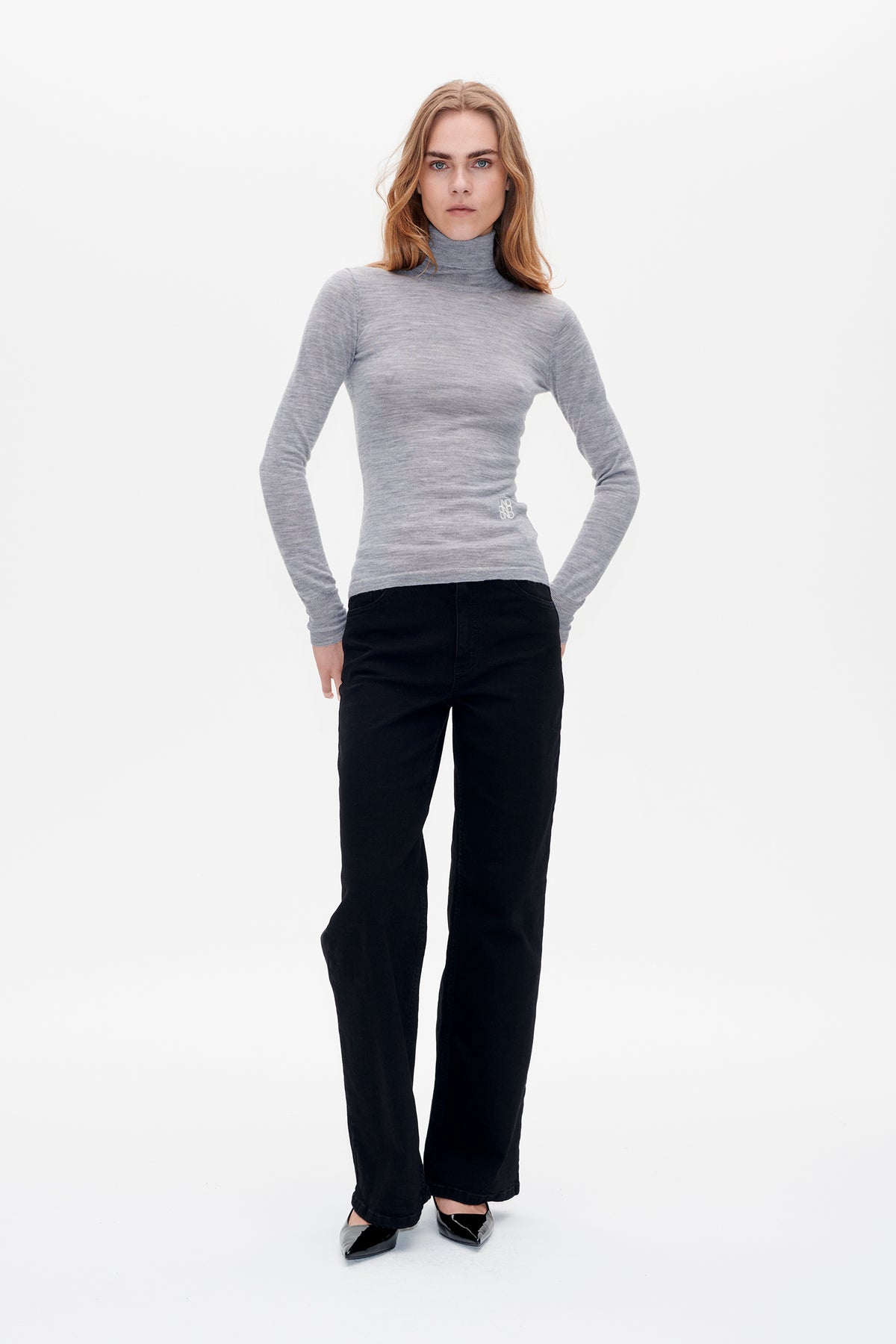 Vooraanzicht van een model dat een getailleerde coltrui met lange mouwen in gemêleerd grijs draagt, met een klein wit taille-embleem, gecombineerd met een zwarte broek met wijde pijpen.