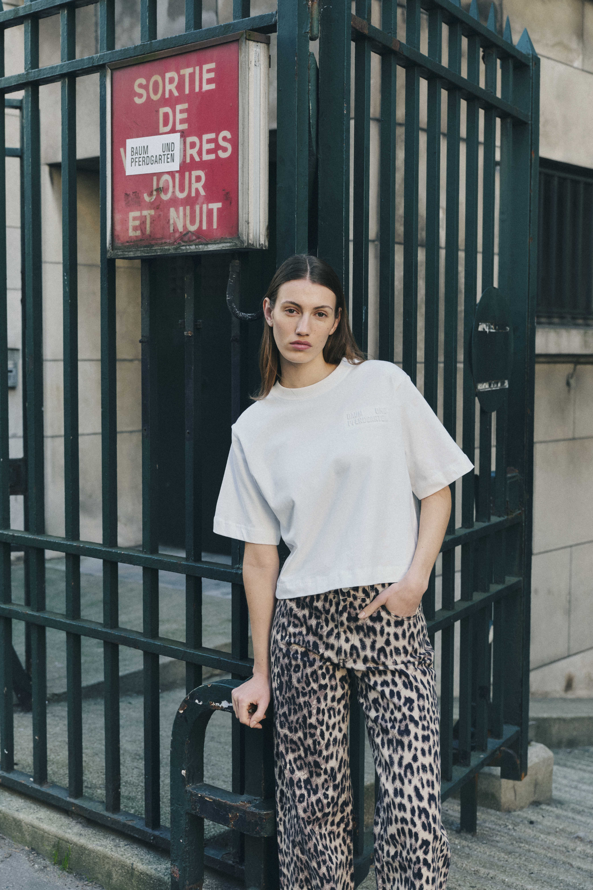 Model wearing an oversized white T-shirt with a small chest logo and beige-and-black leopard-print trousers, leaning against a dark iron gate.
