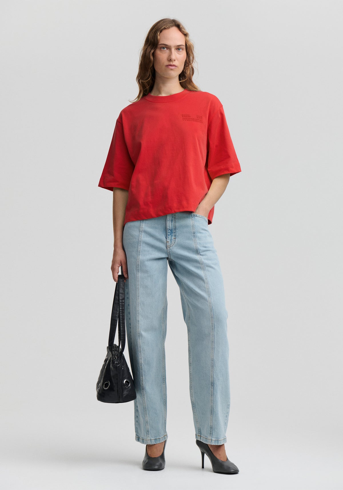 Model wearing an oversized red crew-neck T-shirt with wide short sleeves and tonal chest lettering, light-wash cropped jeans and black heels.
