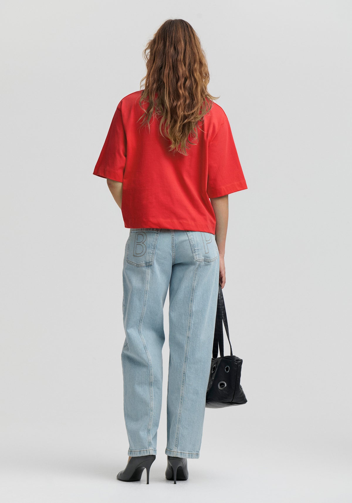 Rear view of a model wearing an oversized bright red short-sleeved T-shirt, light-wash wide-leg jeans with outlined back-pocket lettering, and black heels.