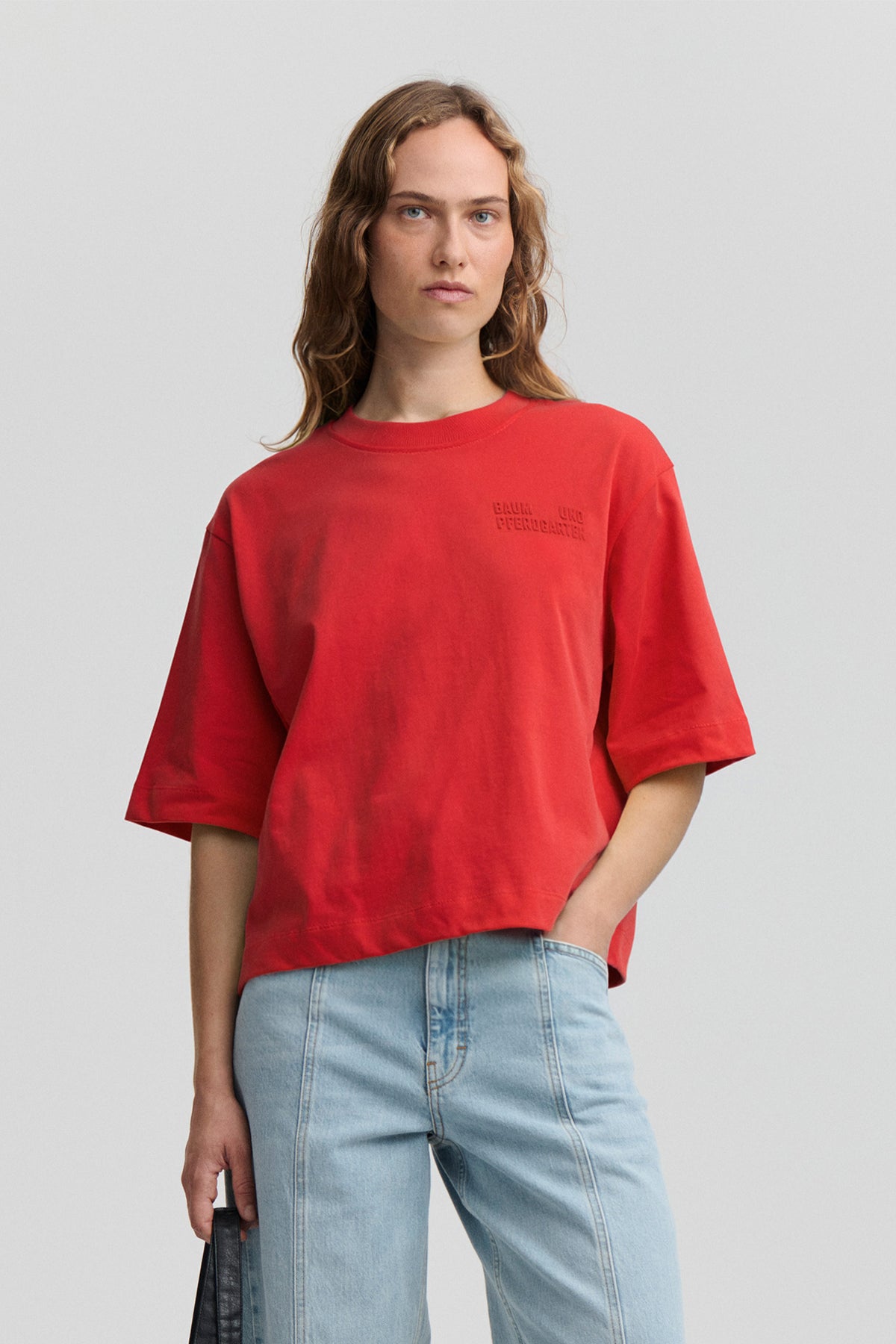 Front view of an oversized red short-sleeve T-shirt with tonal Baum und Pferdgarten lettering on the chest, styled with light-wash jeans.