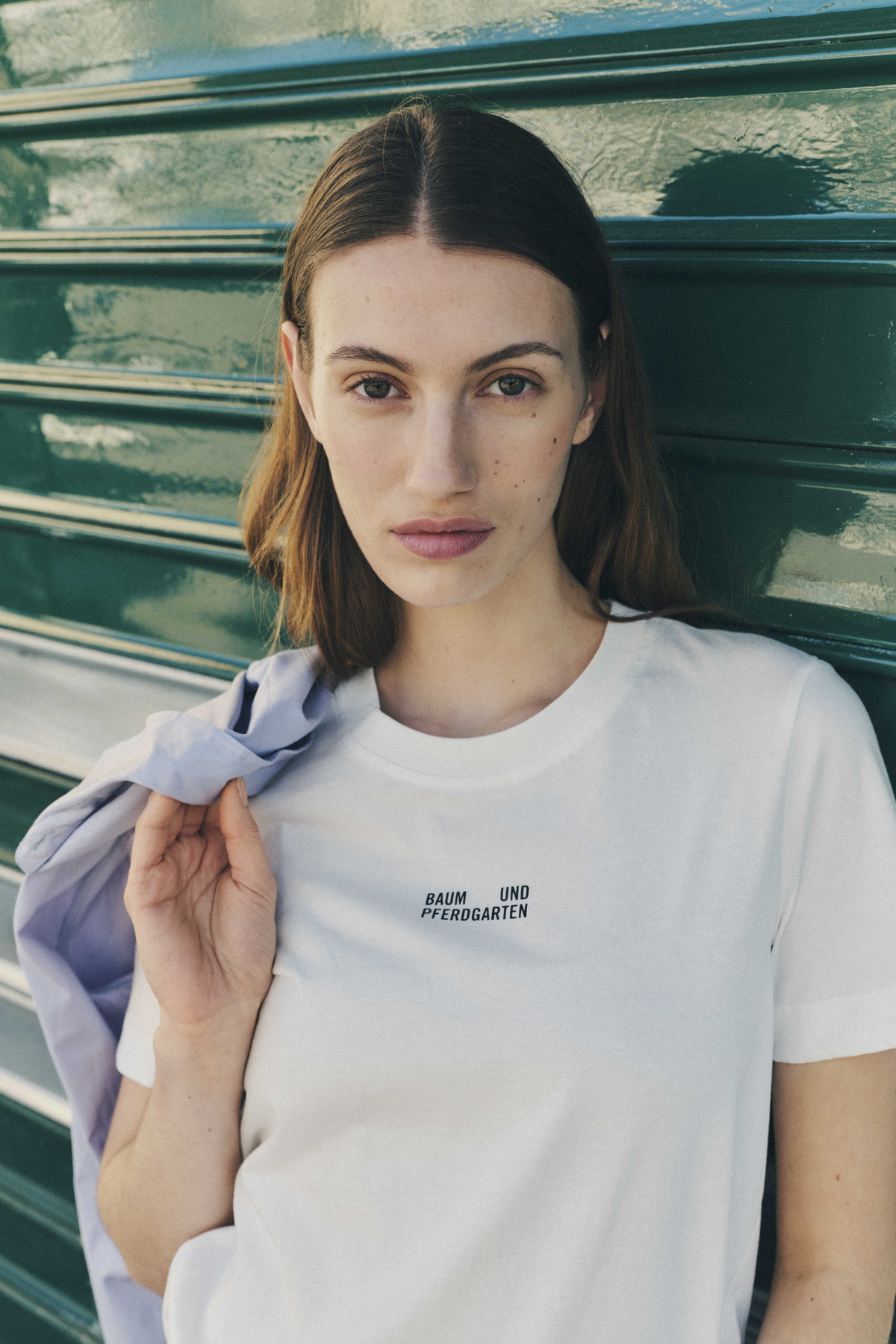 Portrait de campagne en gros plan d'un mannequin portant un T-shirt blanc avec un logo Baum und Pferdgarten sur la poitrine, tenant un vêtement lavande pâle.
