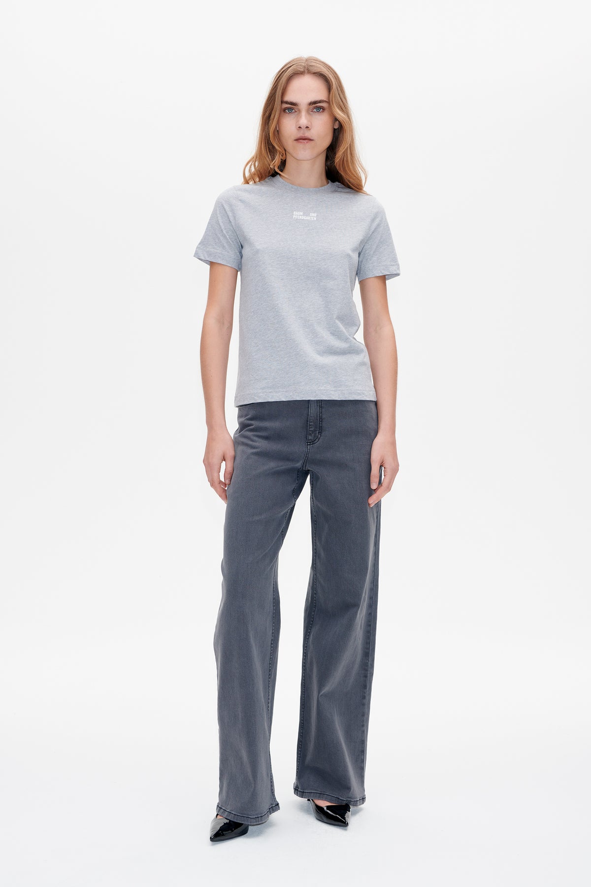 Model trägt ein hellgraues, kurzärmeliges T‑Shirt mit kleinem weißen Brustlogo und dunkelgraue, weitgeschnittene Jeans, Vorderansicht.