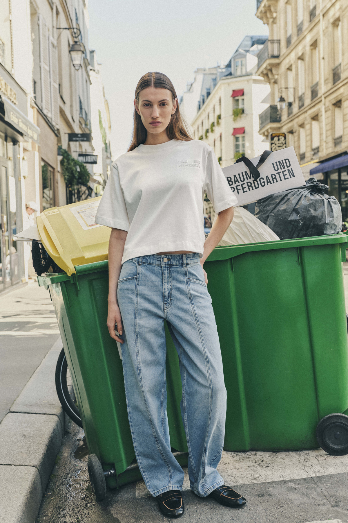 Model wears a white short-sleeved T-shirt and light-wash wide-leg jeans beside green street bins, holding a Baum und Pferdgarten shopping bag.