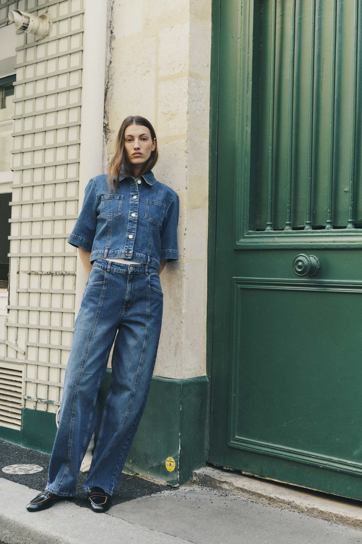 Model wearing a blue denim short-sleeved button-front shirt with chest pockets and matching wide-leg jeans, posed beside a dark green door.