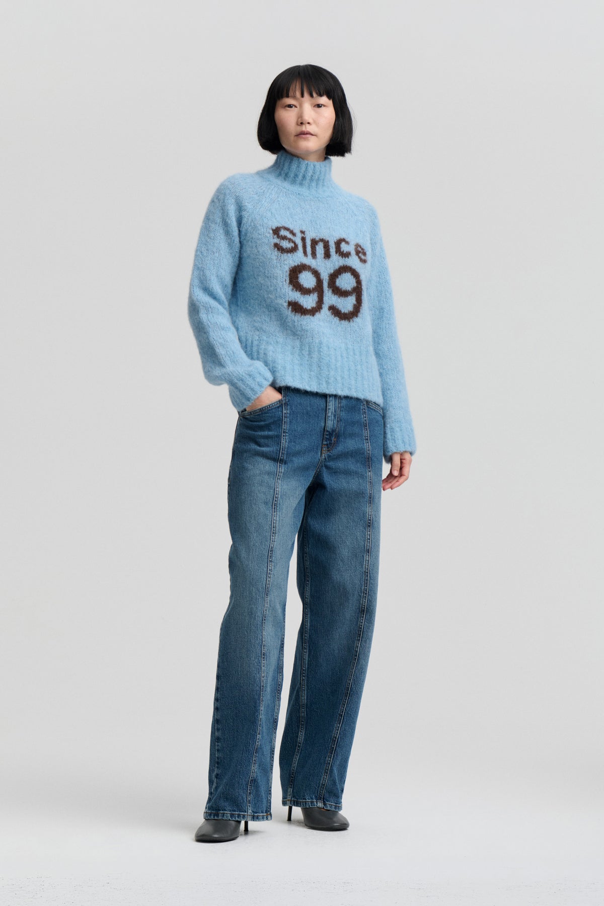 Model wearing a light blue high-neck sweater with brown “Since 99” lettering and blue wide-leg jeans against a plain studio backdrop.