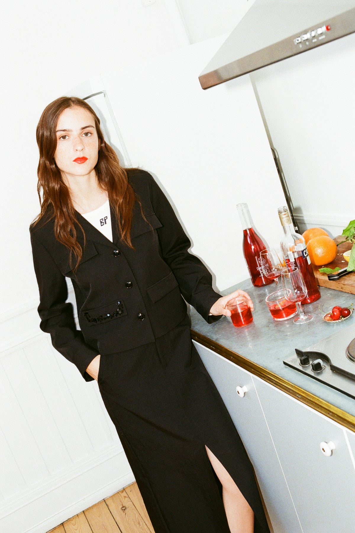 Campaign portrait of a model in a black button-front blazer dress over a white graphic T-shirt, holding a glass at a kitchen counter.