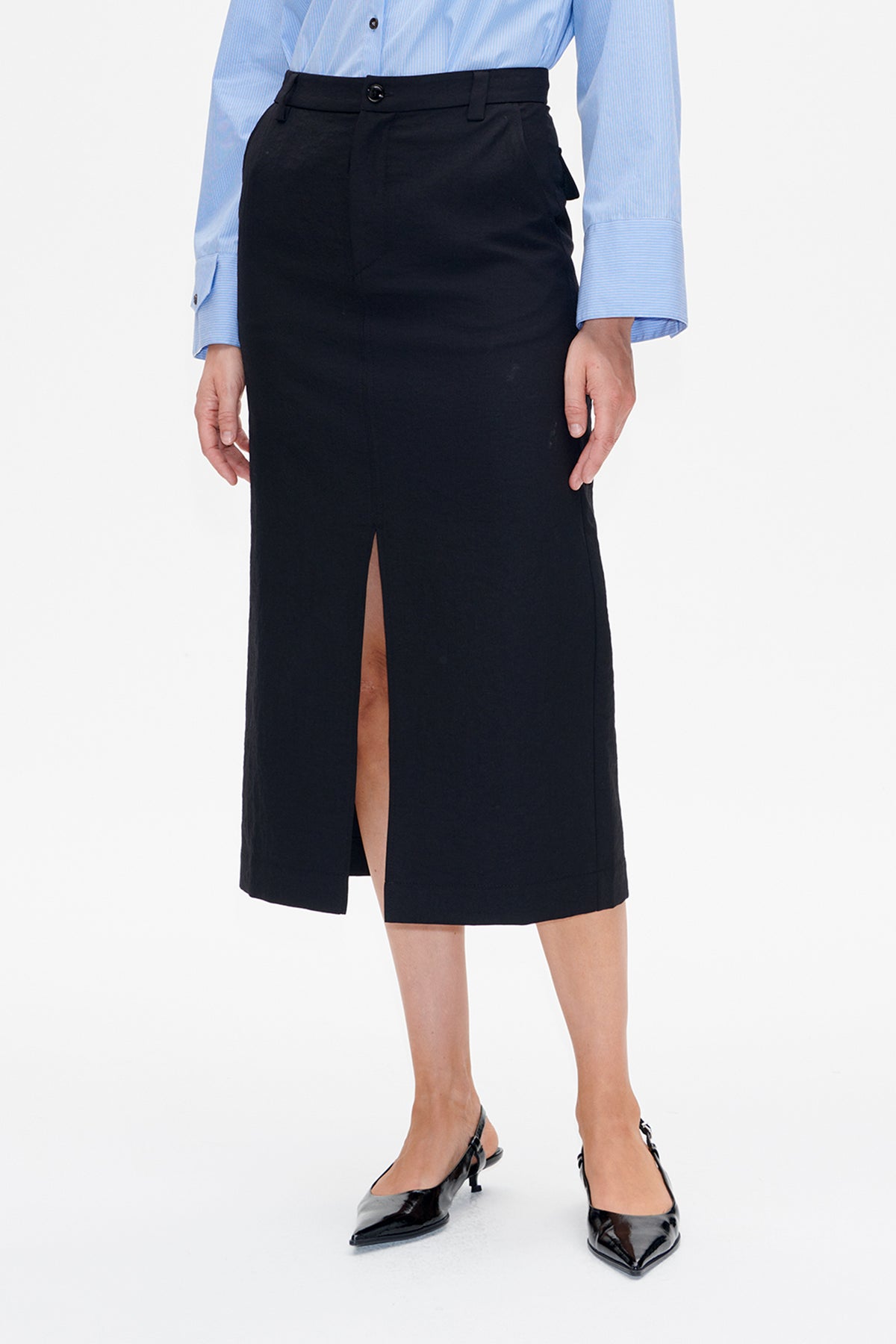 Vooraanzicht van een zwarte midi-rok met hoge taille, ceintuurlussen en een lange voorsplit, gestyled met een blauwe blouse met fijne krijtstrepen.