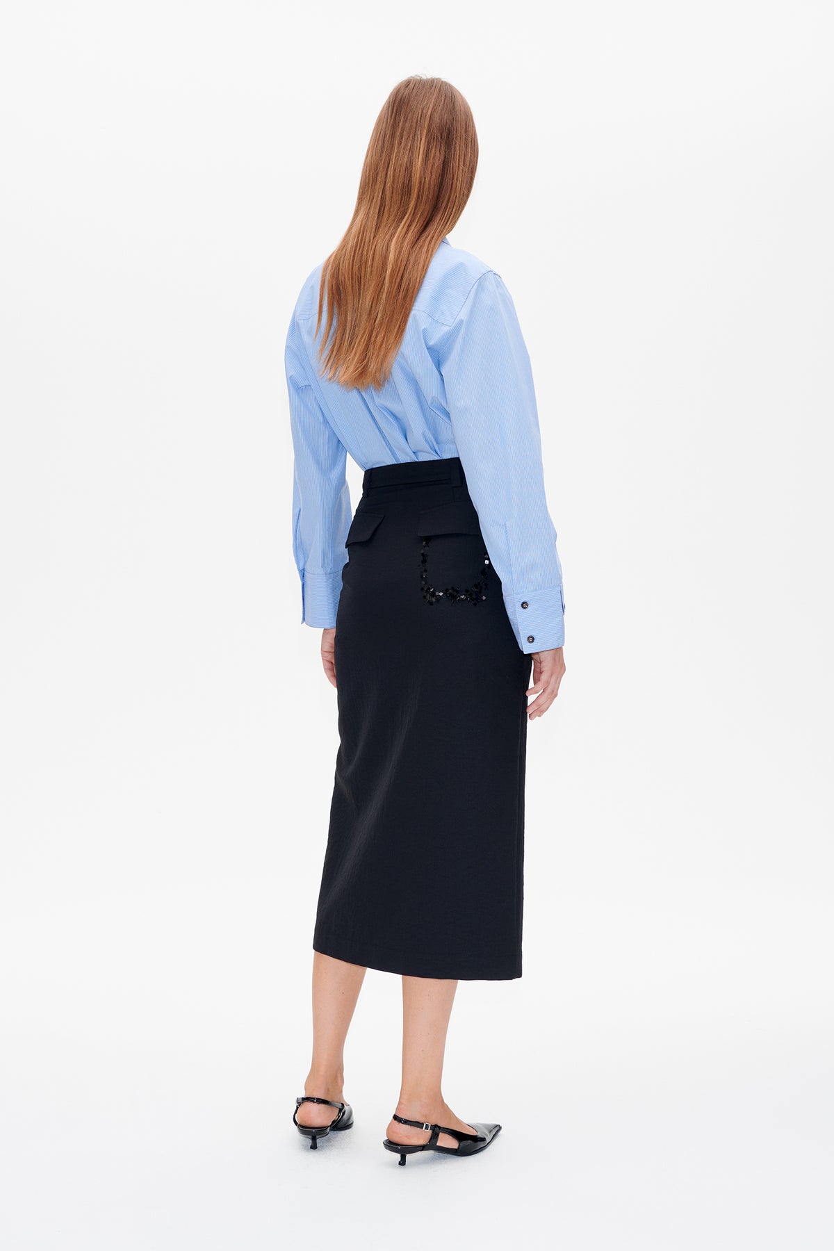 Achteraanzicht van een model in een lichtblauwe, fijn gestreepte blouse en een zwarte midi rok met versierde klepjes op de achterzakken.