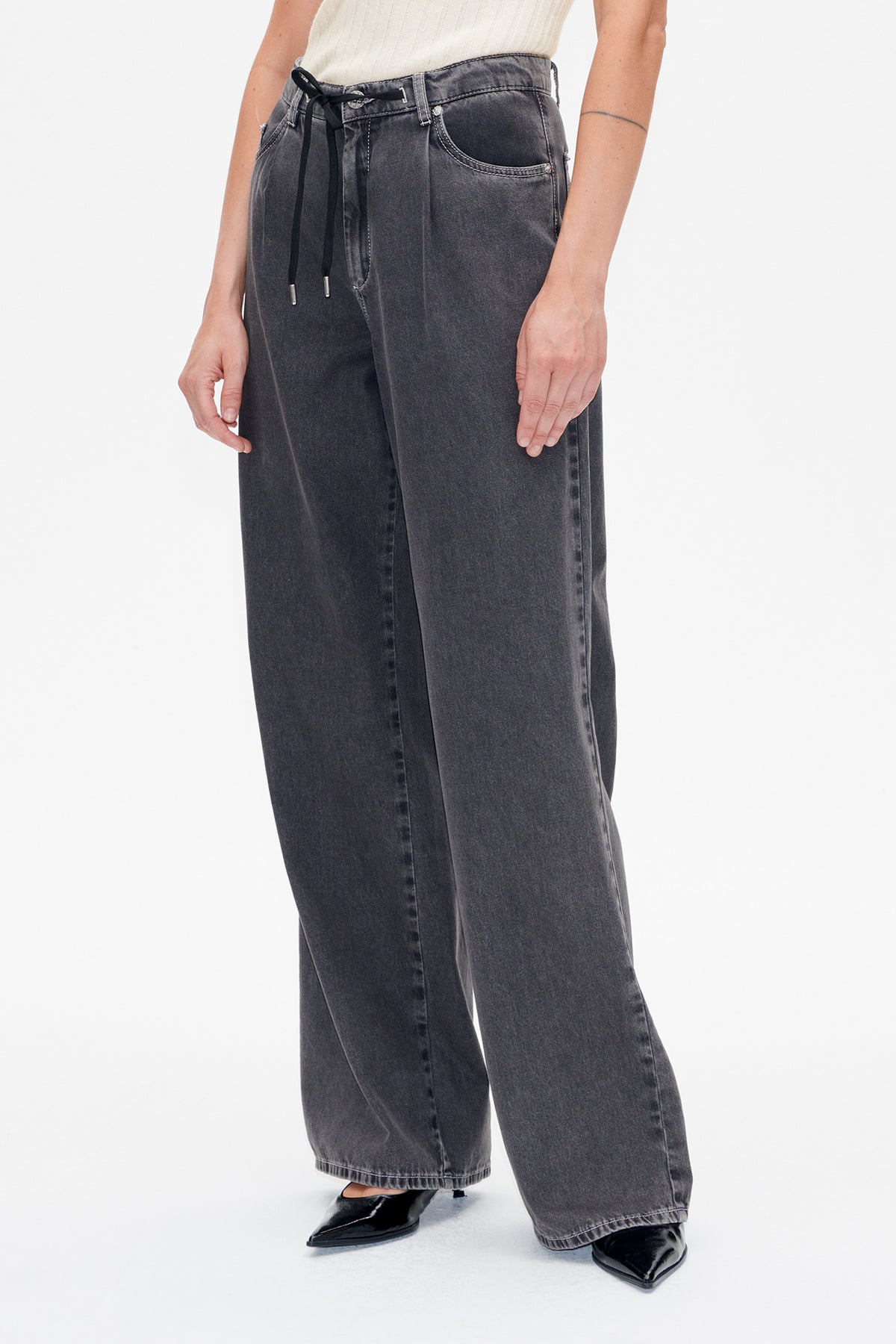 Detail van de voorkant van een wijd uitlopende jeans in antraciet met een vervaagde wassing, voorzakken en een zwarte taille met trekkoord.