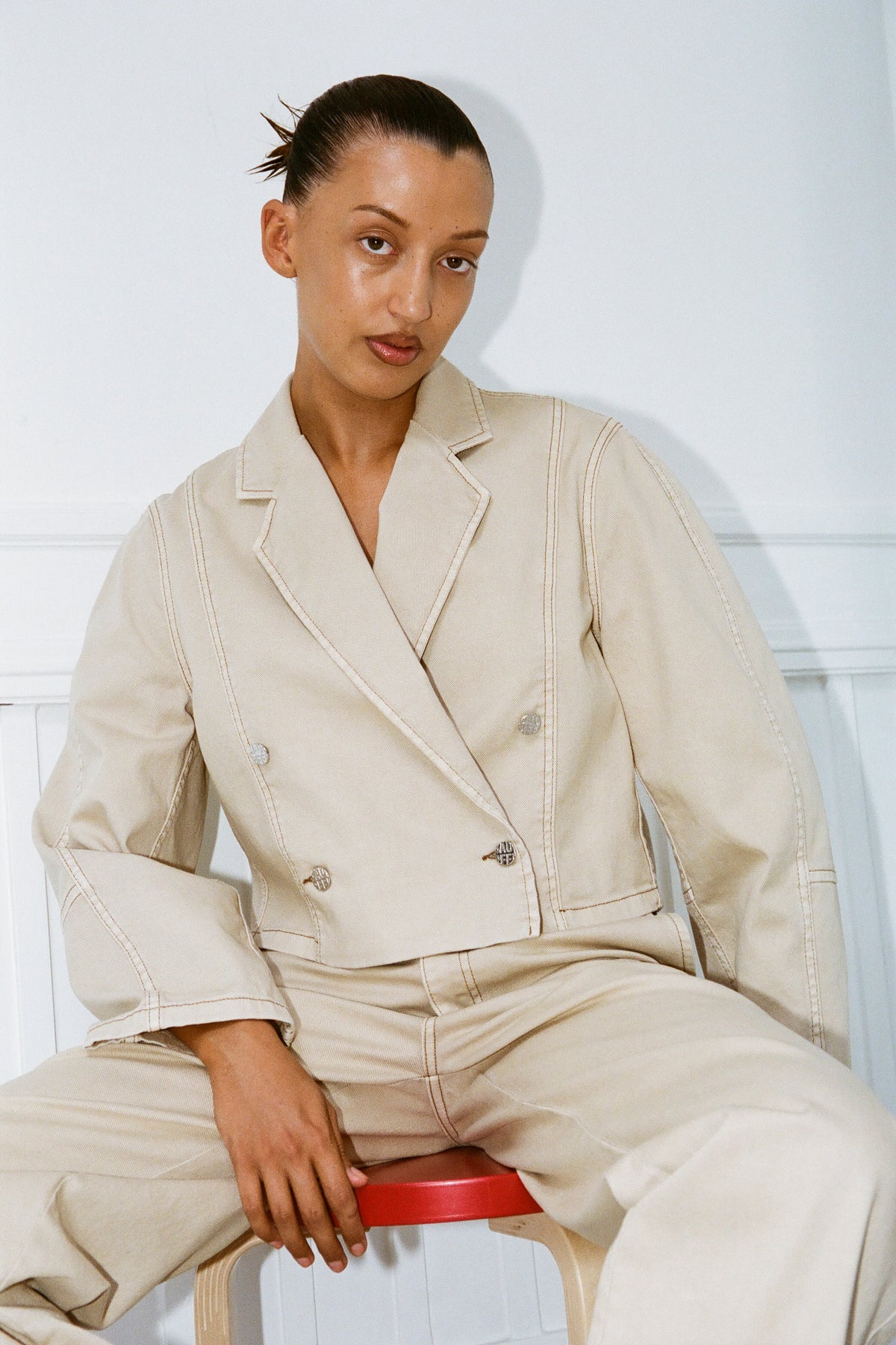 Seated model wearing a pale beige double-breasted jacket with contrast stitching and matching trousers, holding a red book.