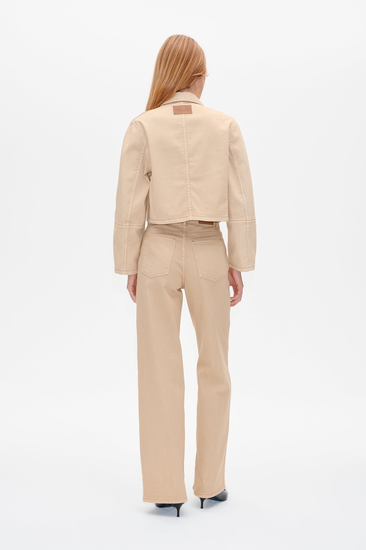 Back view of a model wearing a cropped beige jacket with seam detailing and matching wide-leg trousers.