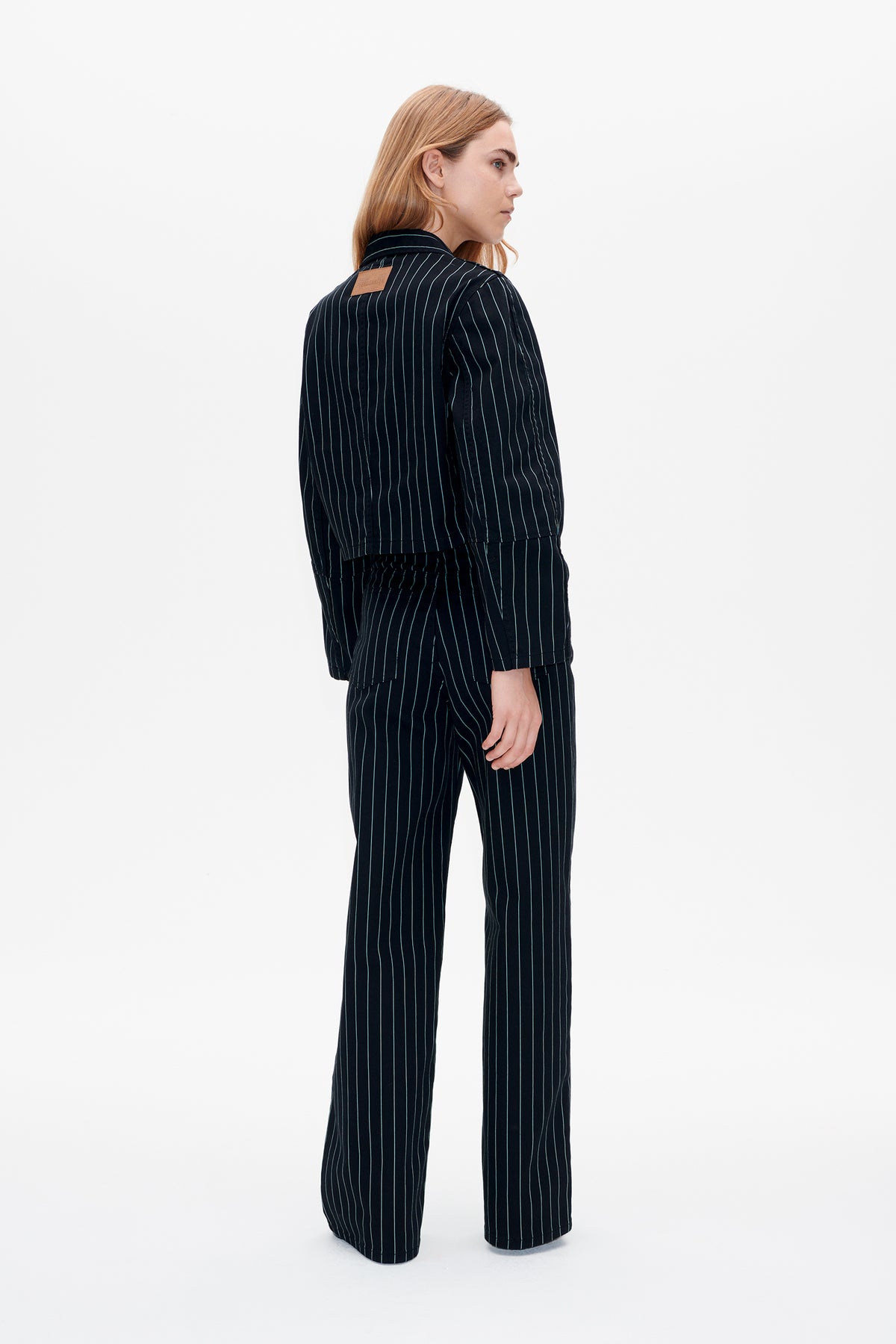 Rear three-quarter view of a model wearing a dark navy pinstripe jacket and matching wide-leg trousers, with a tan patch at the jacket collar.