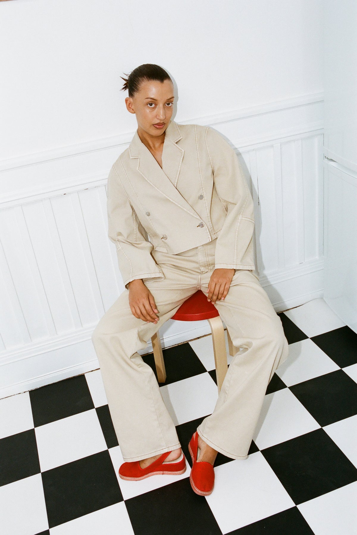 Model seated against a white paneled wall in a cream contrast-stitched jacket and wide-leg trousers with bright red shoes.