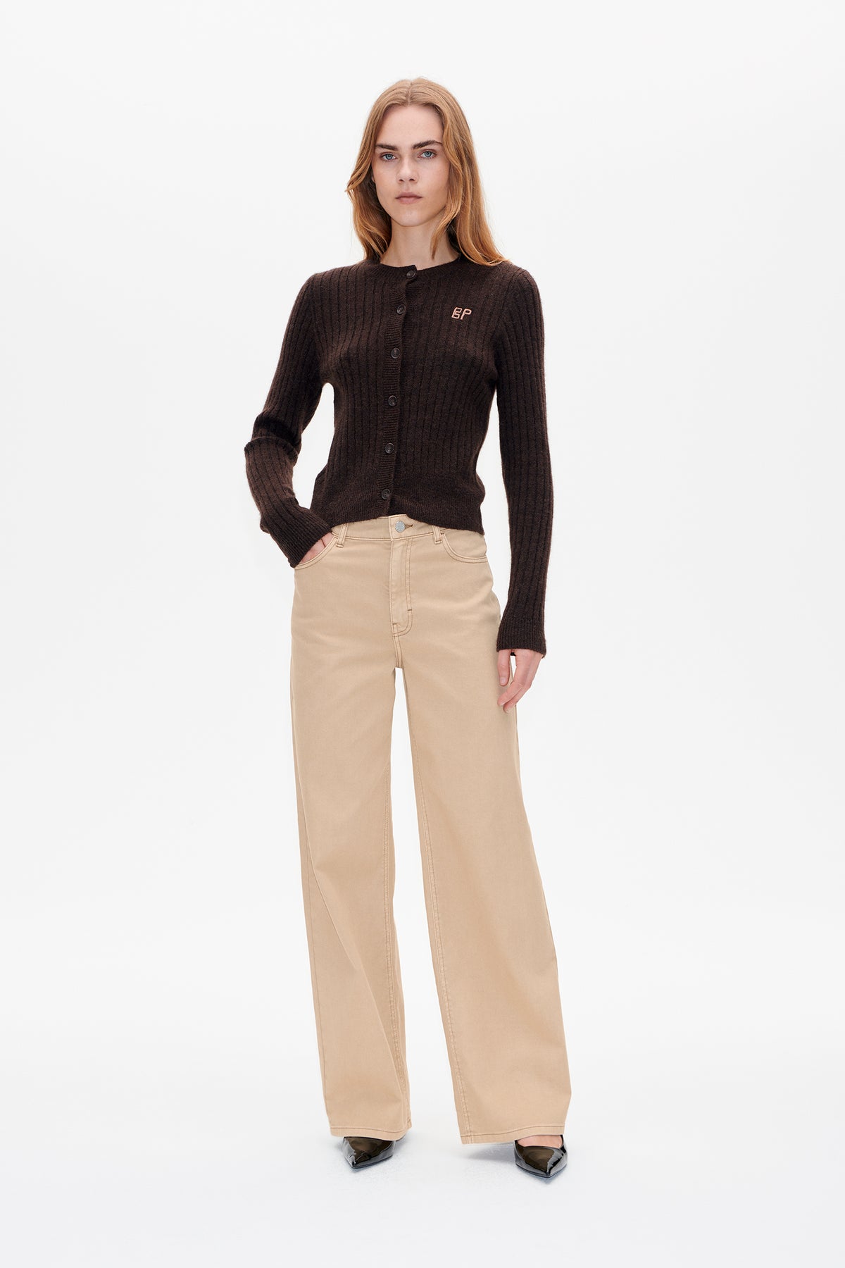 Vorderansicht eines Models in einem dunkelbraunen, gerippten Cardigan mit Knopfleiste und kleinem Brustlogo, beigen, weit geschnittenen Hosen und schwarzen, spitzen Schuhen.