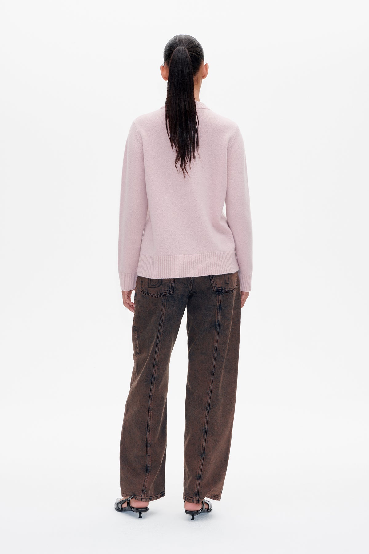 Back view of a model wearing a pale pink long-sleeved sweater with ribbed cuffs and hem, brown washed jeans and black sandals.