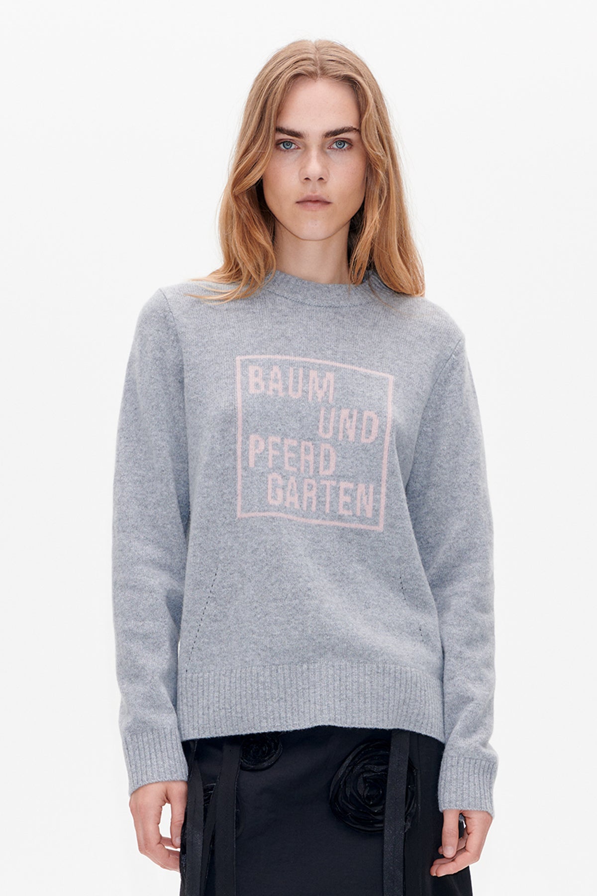 Front view of a model wearing a heather-grey crew-neck sweater with a pale pink BAUM UND PFERDGARTEN graphic and a black skirt.