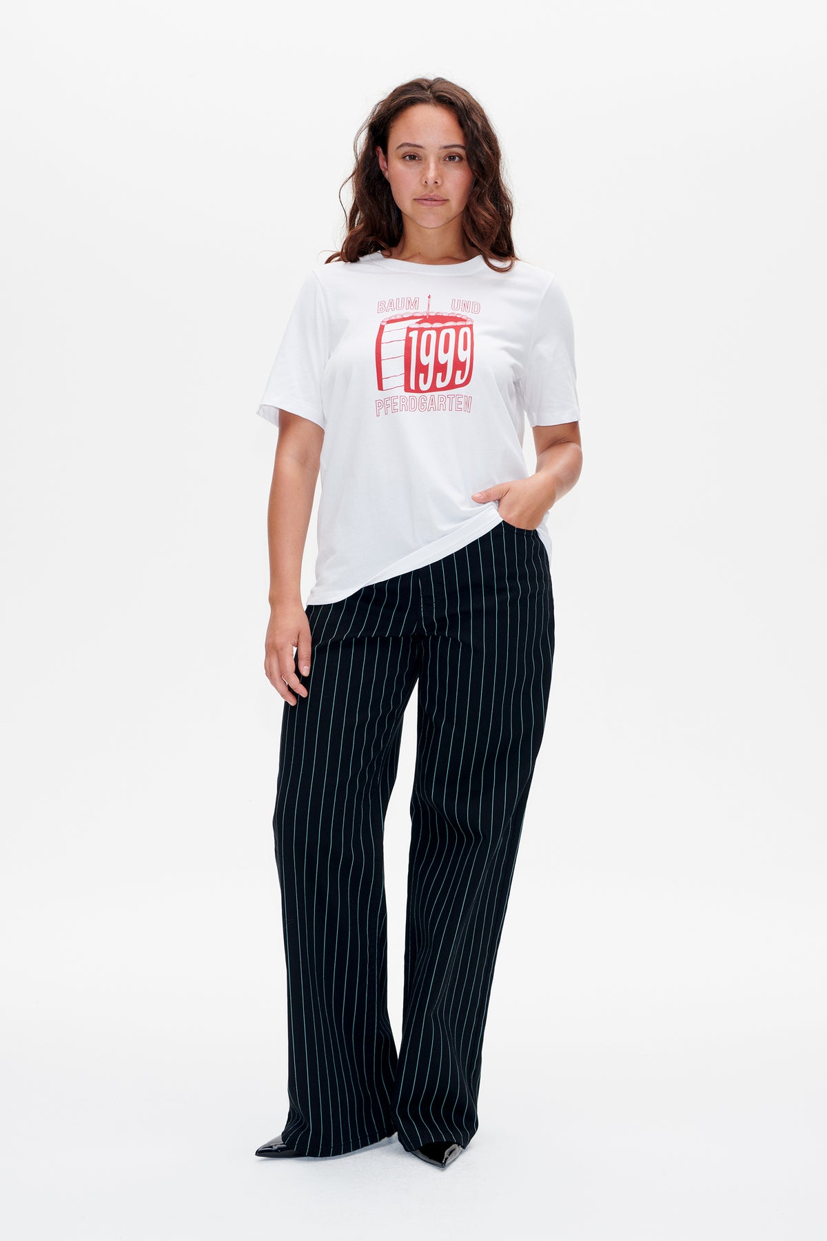 Das Model trägt ein weißes T-Shirt mit roter 1999-Tortengrafik und eine schwarze Hose mit feinen vertikalen Streifen.