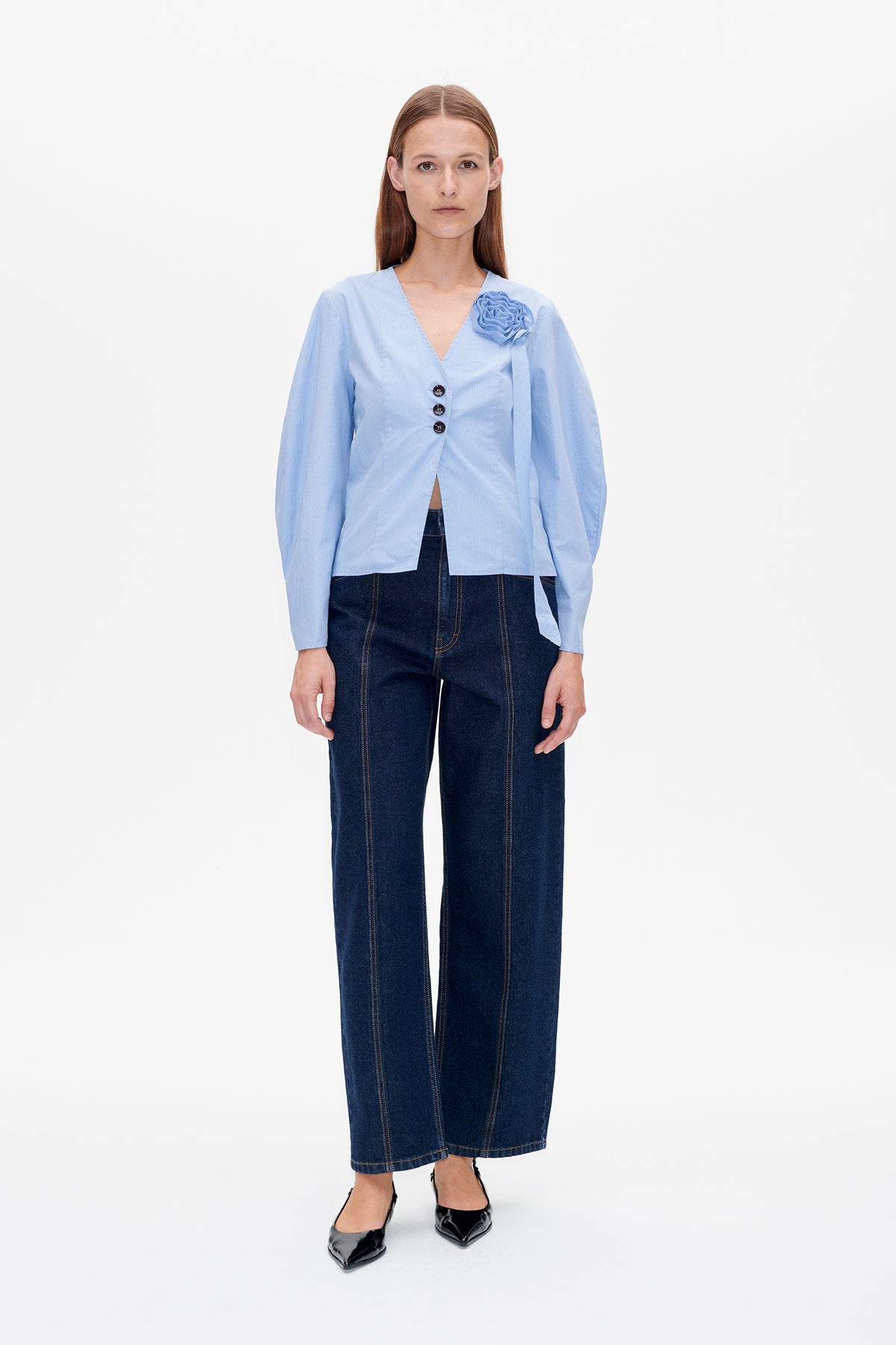 Vorderansicht einer hellblauen Bluse mit Nadelstreifen, tiefem V-Ausschnitt, drei Knöpfen und Stoffblume als Detail, kombiniert mit dunklen Jeans mit weitem Bein.