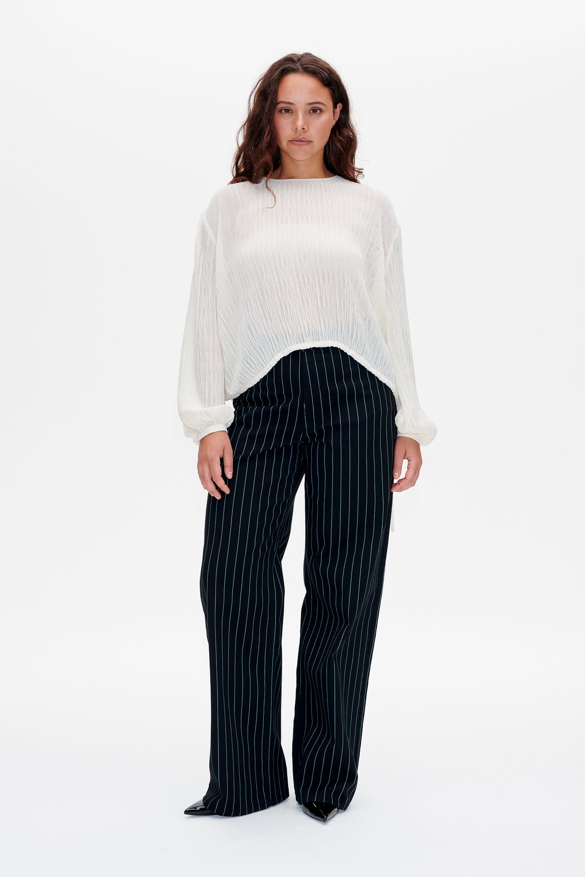 Vooraanzicht van een model in een witte, gestructureerde blouse met lange, geplooide mouwen en een marineblauwe broek met wijde pijpen en fijne verticale krijtstrepen.
