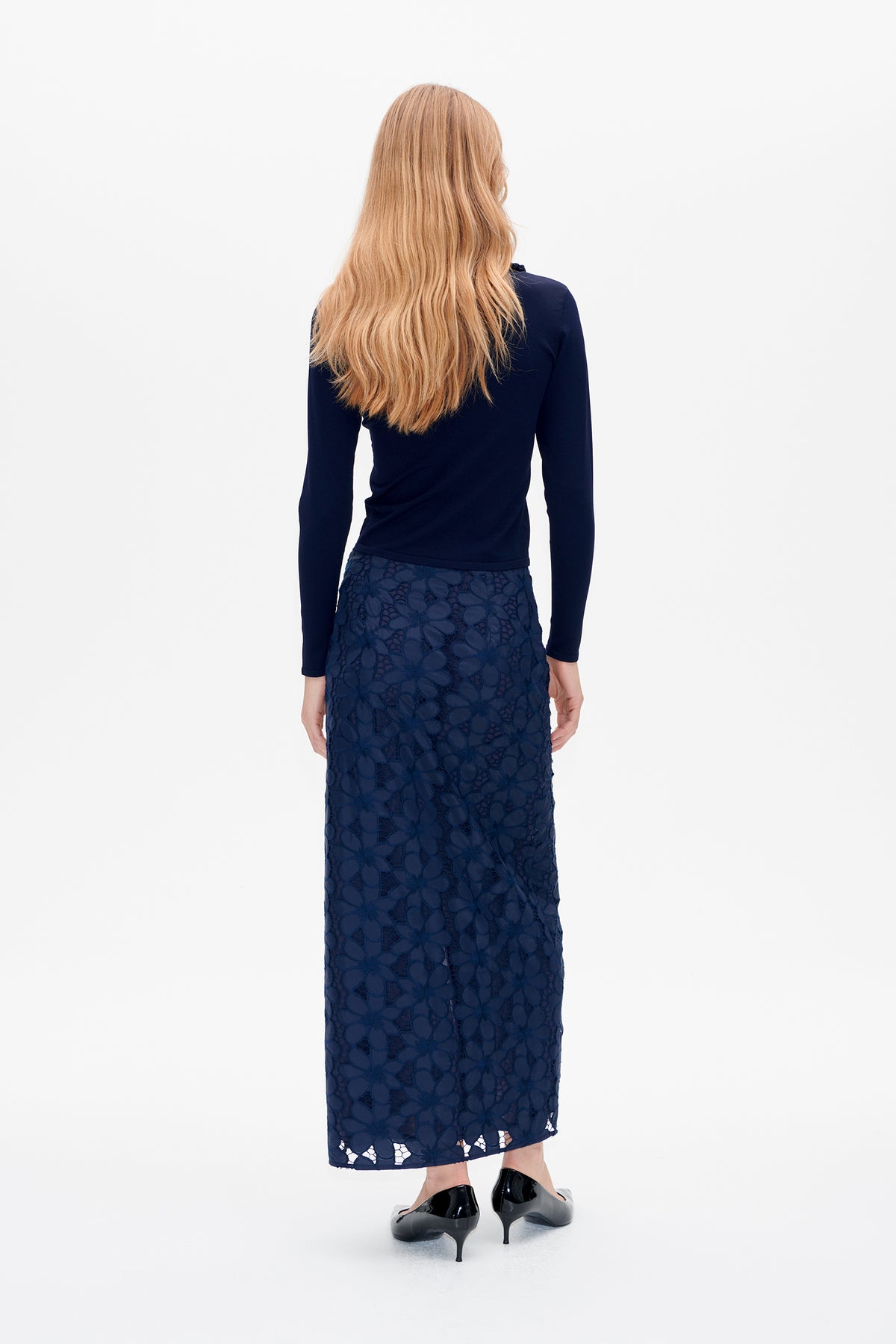 Achteraanzicht van een model dat een getailleerde marineblauwe top met lange mouwen en een marineblauwe kanten rok met bloemen tot op de enkel draagt.