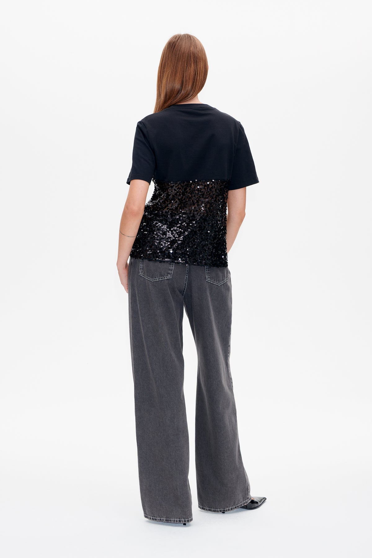 Back view of a black short-sleeved T-shirt with a sparkling sequin panel across the lower back, paired with charcoal wide-leg jeans.